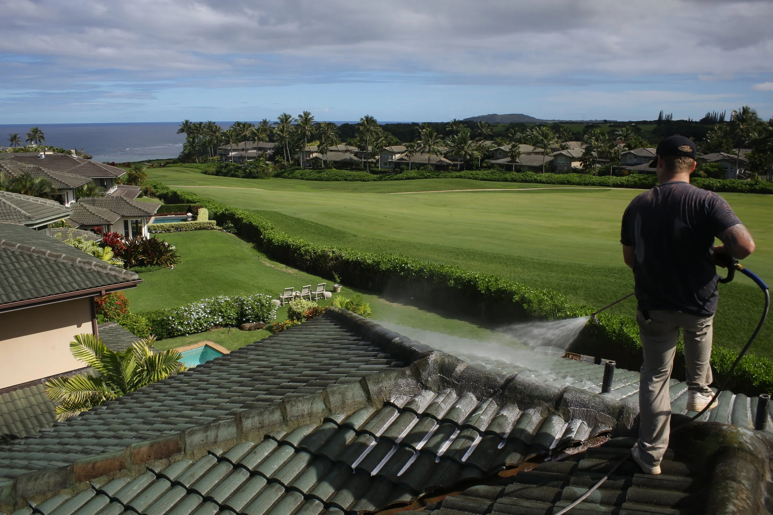Man pressure washing a tiled house roof with a lush green golf course, palm trees, and ocean in the background.