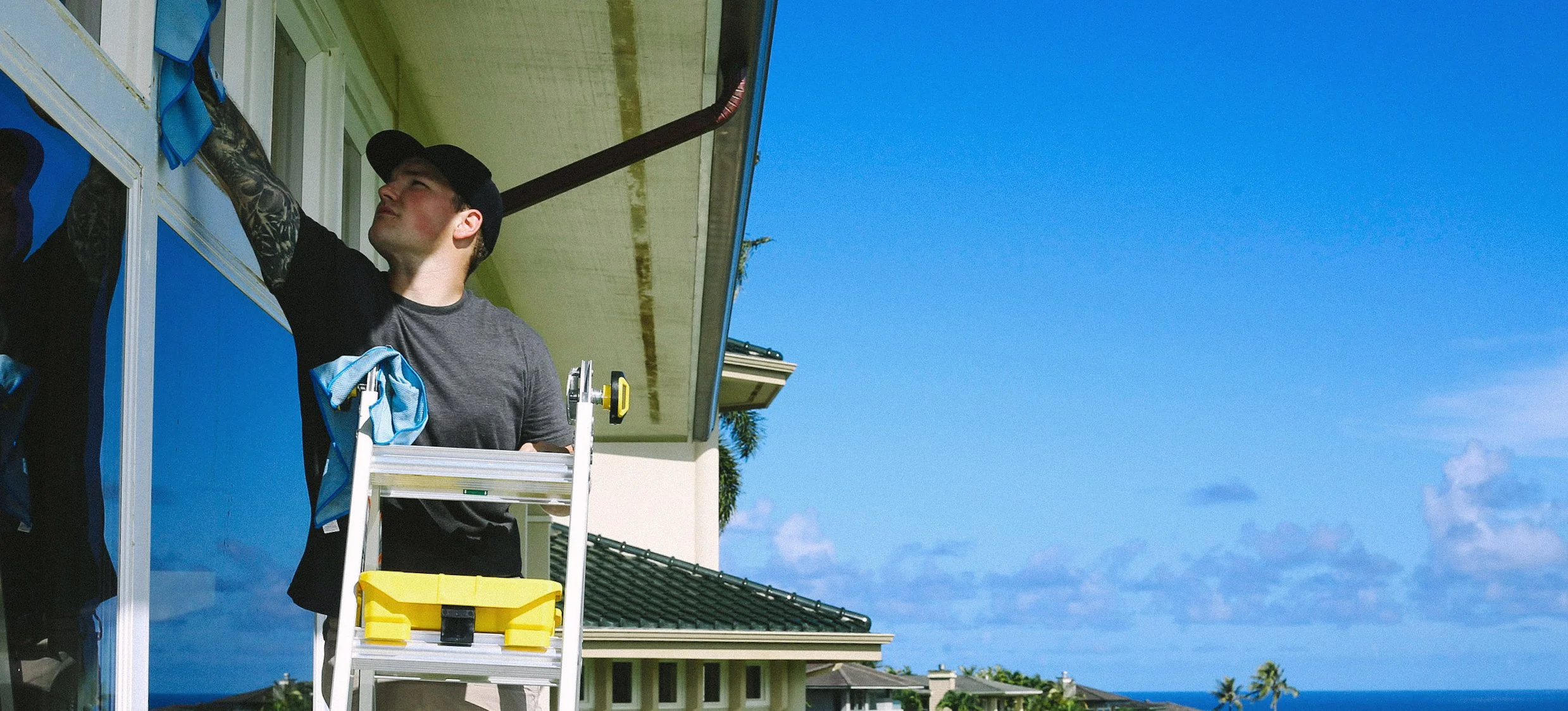 A man is on a ladder cleaning windows outside a house under a clear blue sky, with houses and palm trees visible in the background.