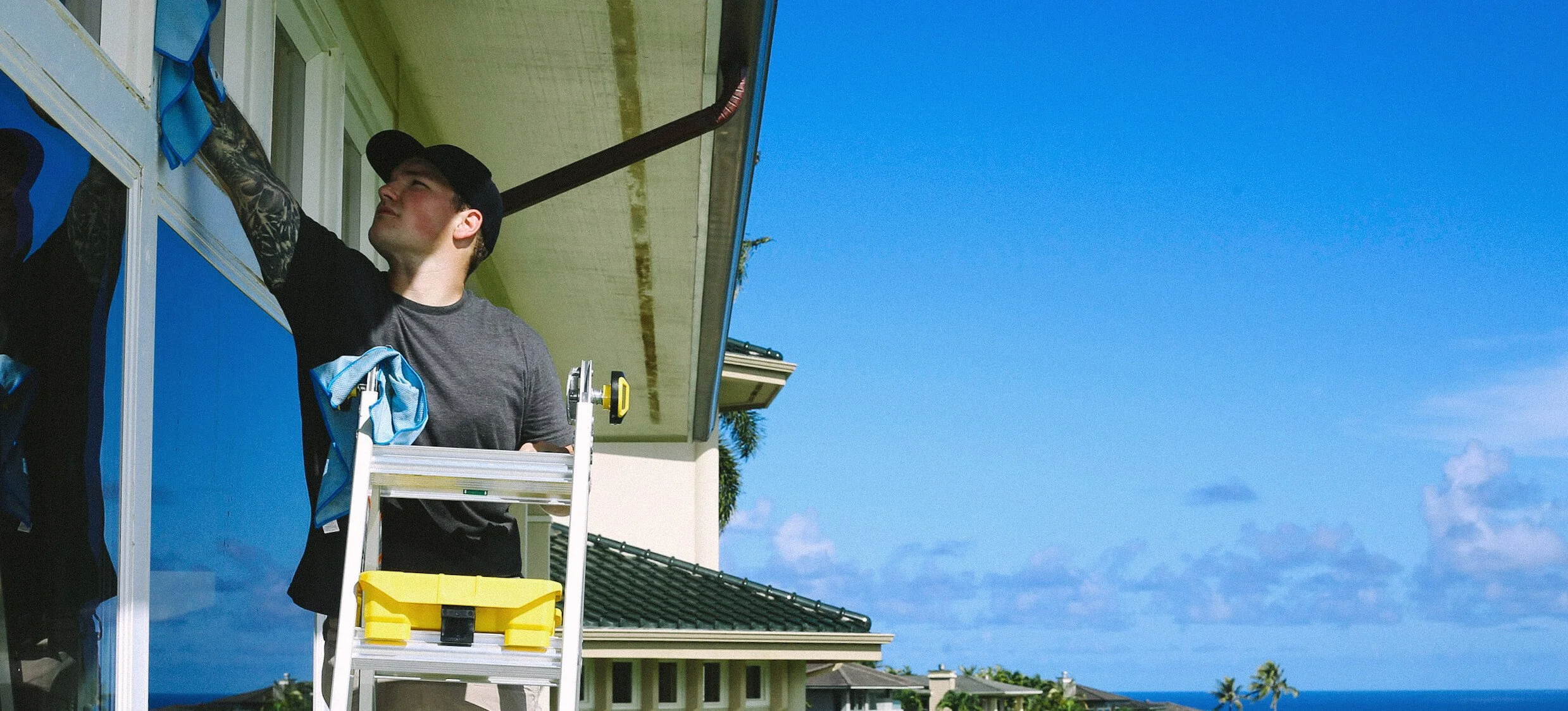 A man wearing a black cap and gray shirt standing on a ladder and reaching into a window of a house, with a view of the ocean and palm trees in the background under a clear blue sky.