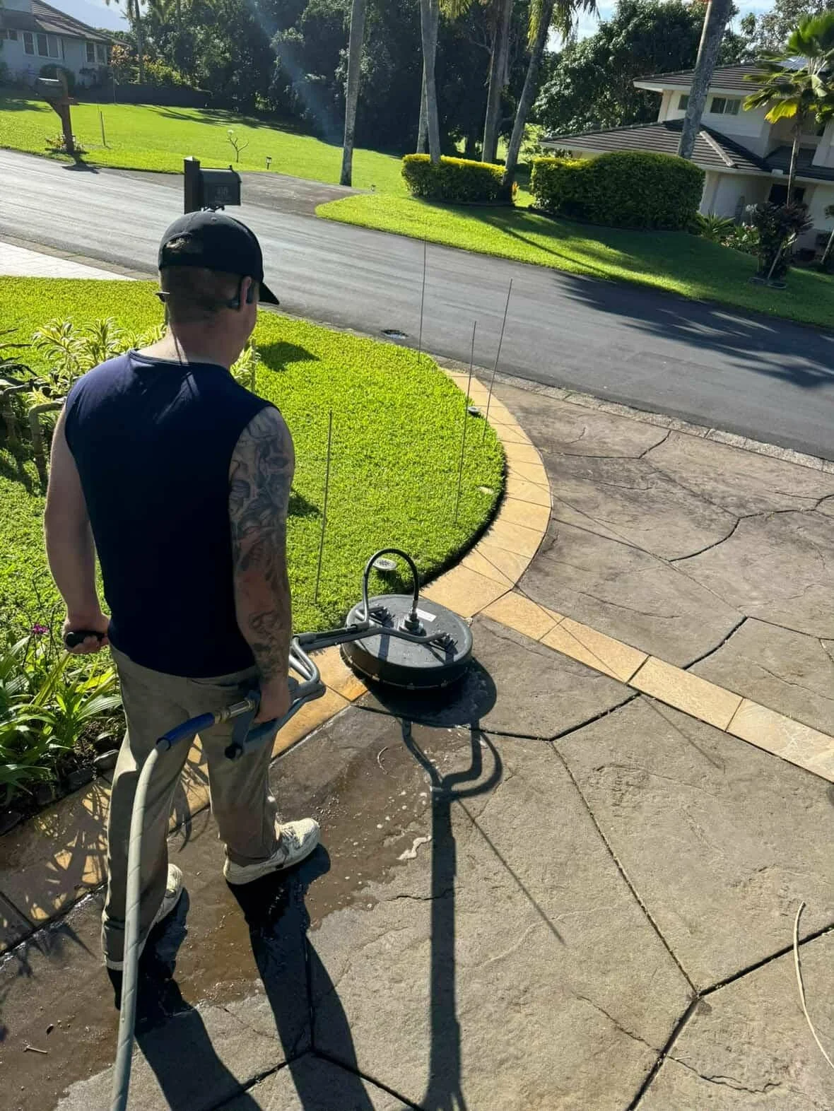 A man wearing a black cap, sunglasses, a sleeveless shirt, beige pants, and white shoes is pressure washing the concrete driveway in front of a house. He is holding a pressure washer wand connected to a machine on the ground, with water spraying on t