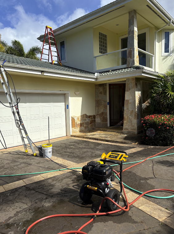 Residential home undergoing exterior work with ladders, hoses, and a pressure washer on the driveway, and a two-story house with a balcony and stone accents.