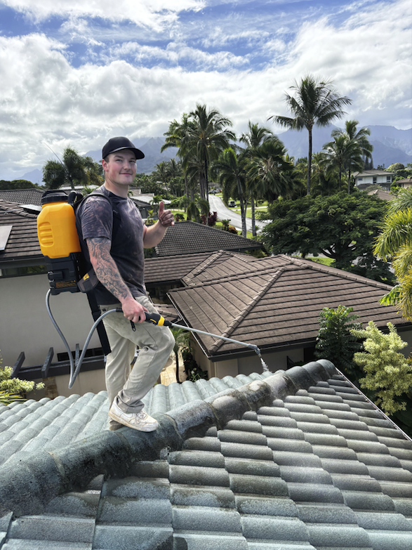 A young person standing on a rooftop, wearing a black cap, a backpack sprayer, beige pants, and sneakers, holding a spray wand, with a background of palm trees, rooftops, and mountains under a partly cloudy sky.