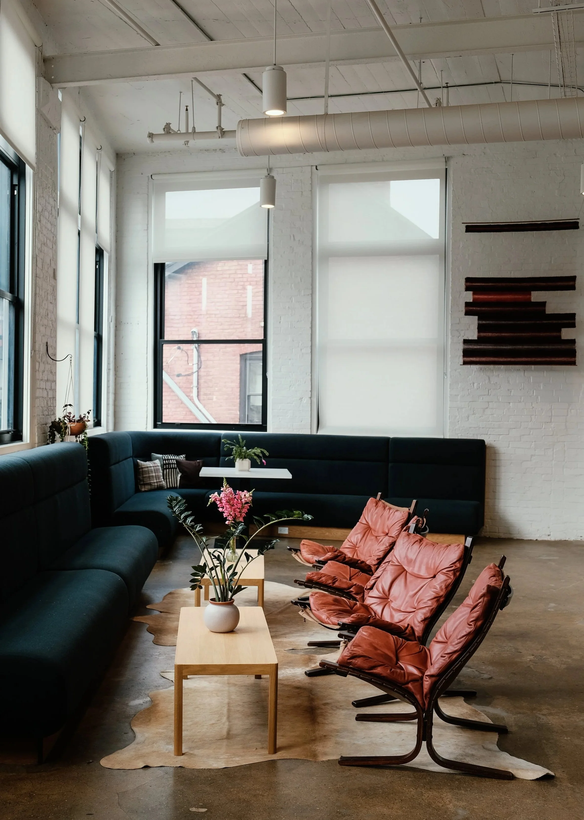 Interior of a modern, spacious co-working or lounge area with large windows, white walls, and eclectic furniture including white and red chairs, a blue sofa, and wooden tables and shelves, decorated with plants and rugs.