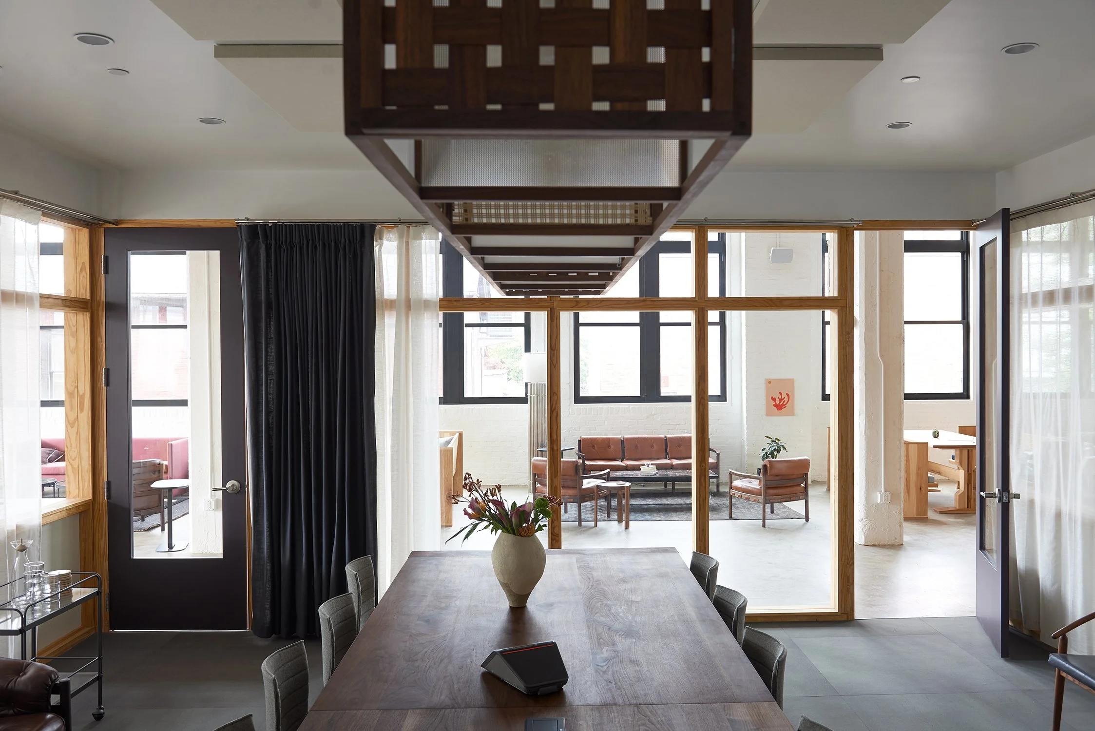 A dining room with a large wooden table, a white vase with flowers, and an overhead wooden shelf; views of a lounge area through open double doors with black frames, featuring sofas and chairs, in a modern space with white walls and windows.