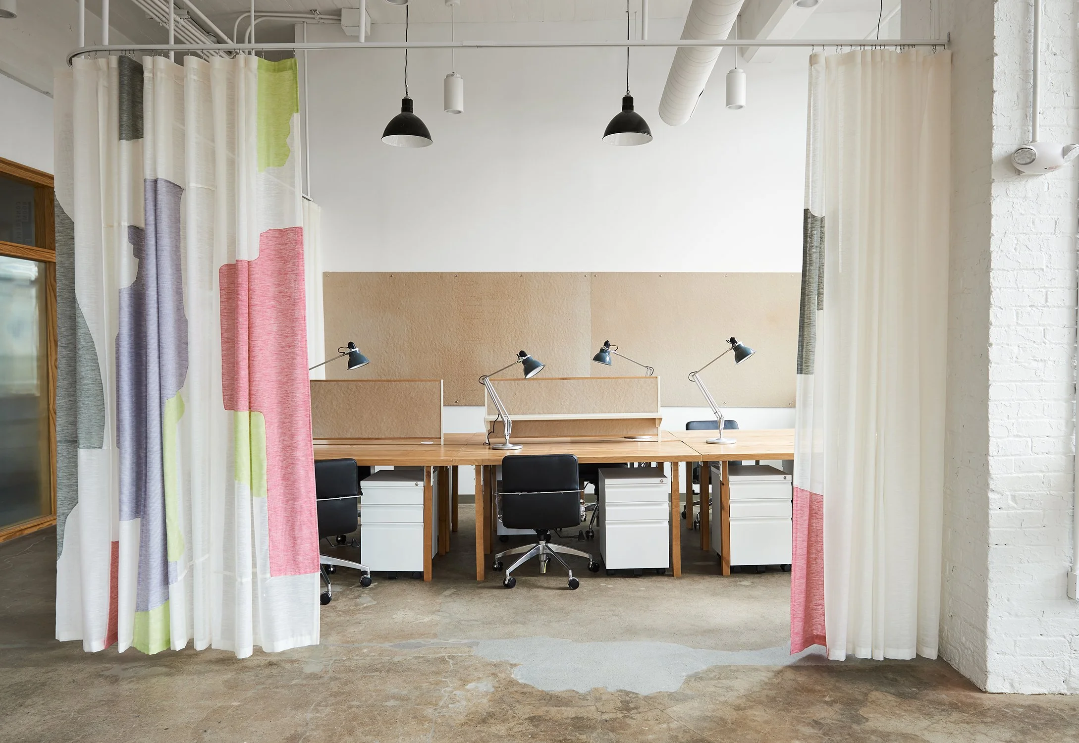 Empty modern office space with four workstations, black chairs, adjustable desk lamps, beige pinboards, and colorful curtains in the foreground.