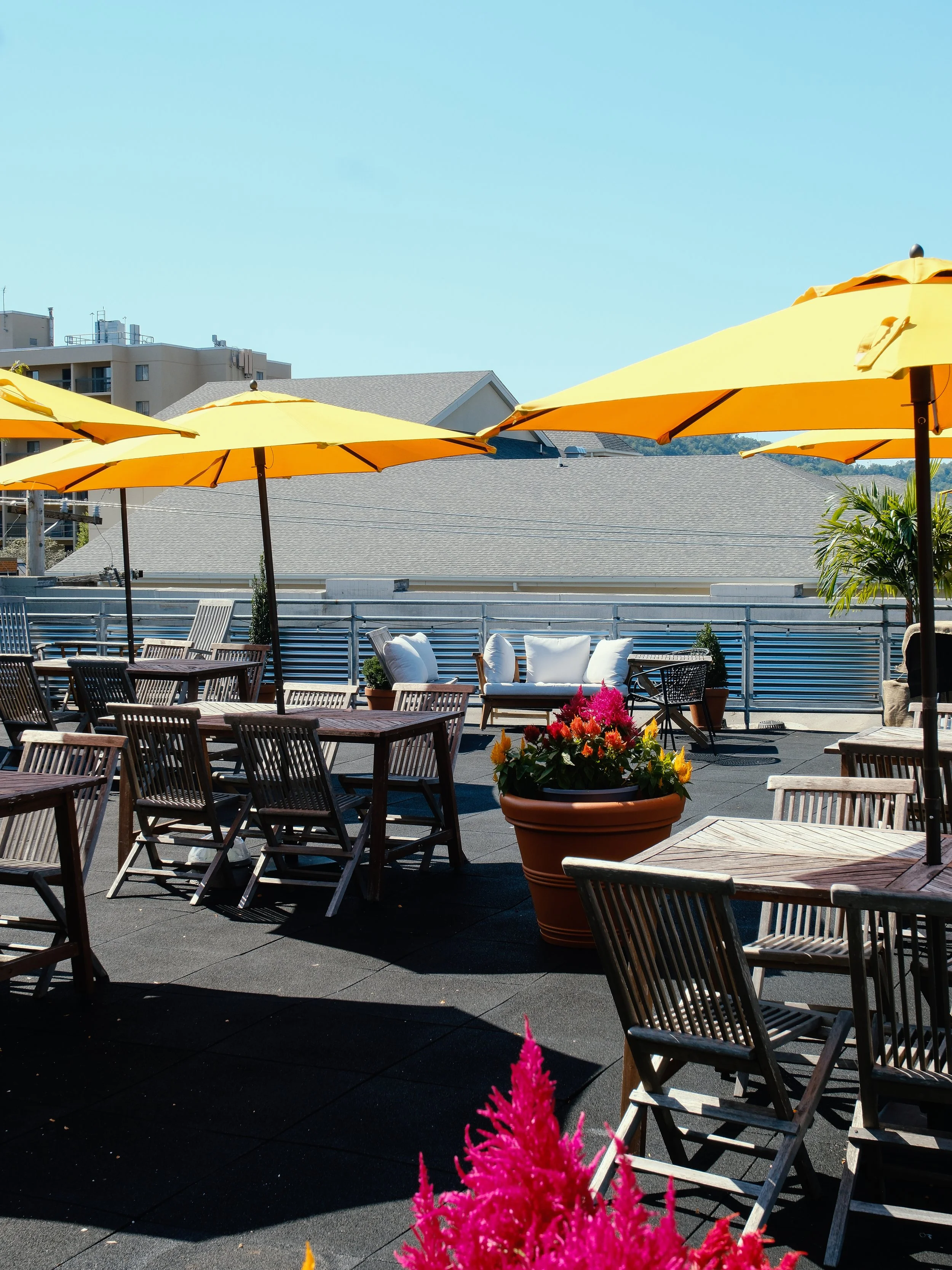 Rooftop patio with yellow umbrella, wooden seating with white cushions, and a city skyline in the background.