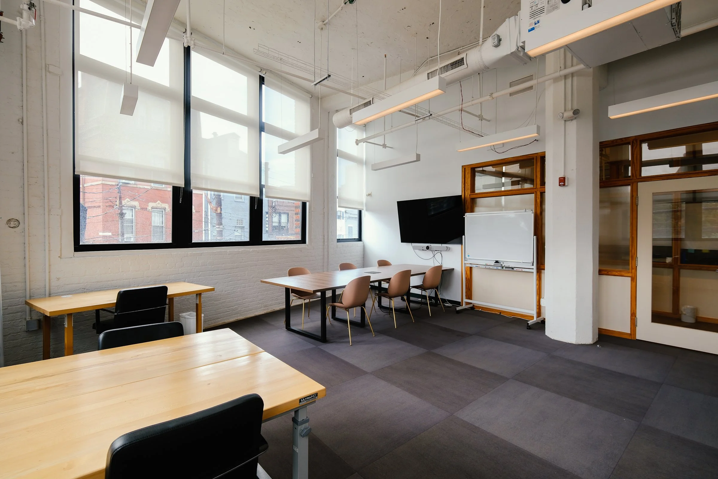 Modern meeting room with large windows, white brick walls, and multiple tables and chairs. Features a large TV and whiteboard on the wall.