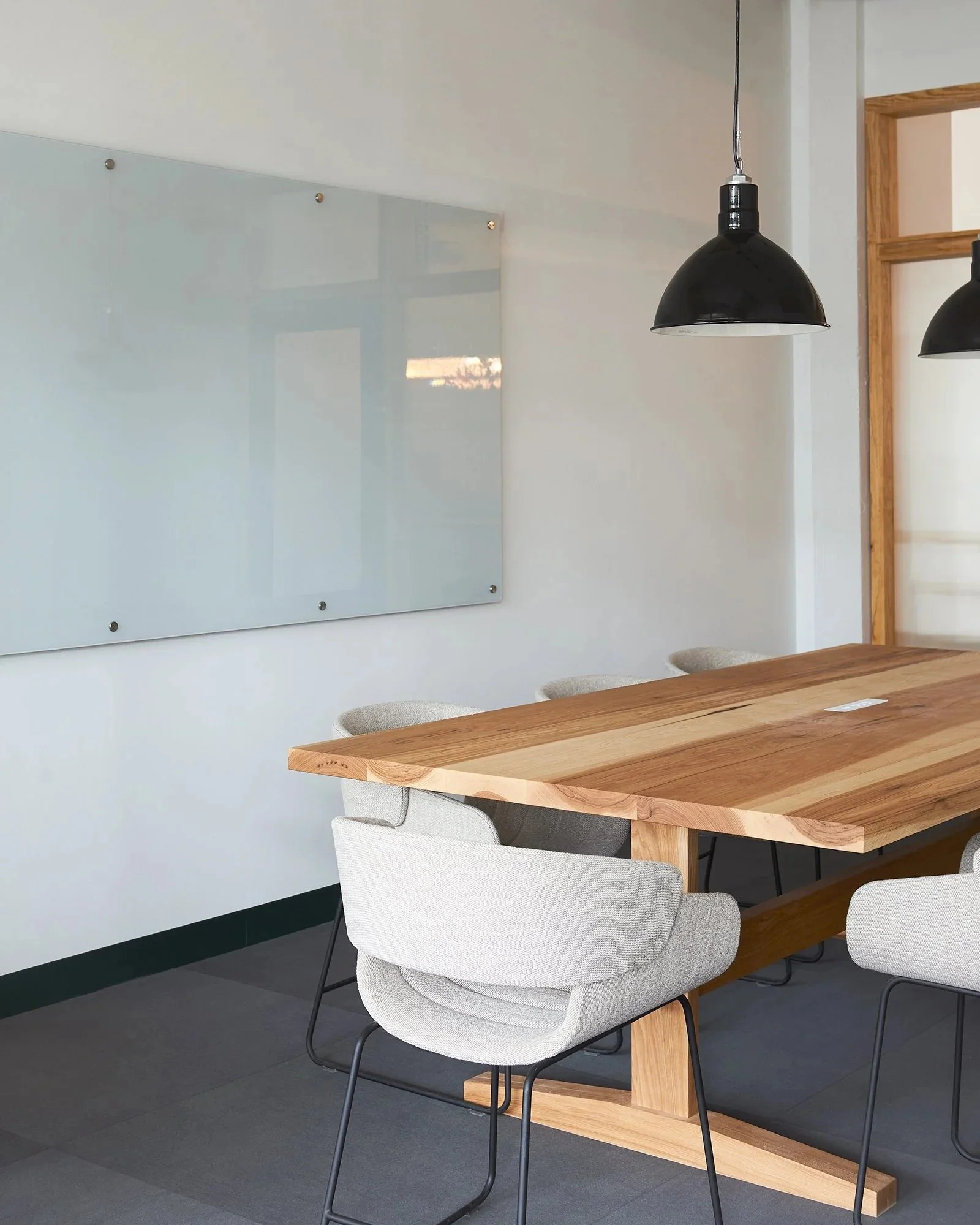 Empty conference room with a large wooden table, eight upholstered chairs, hanging pendant lights, a potted plant, and a glass door leading to another room.