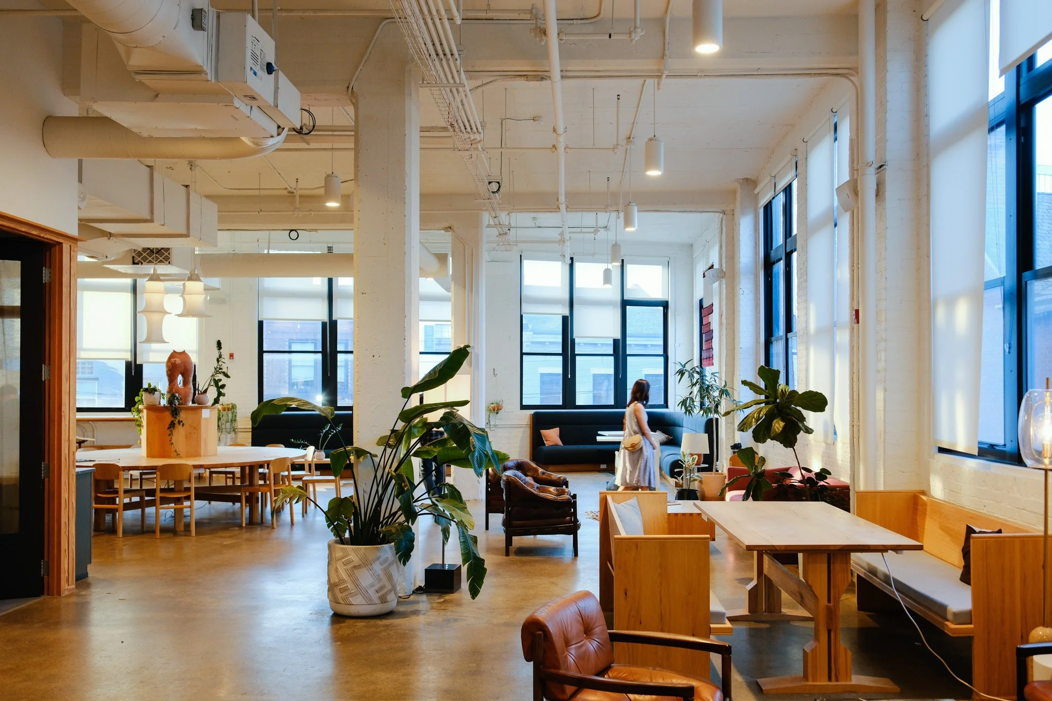 Interior of a modern, bright cafe with large windows, wooden and leather furniture, and potted plants. A woman is walking in the background.