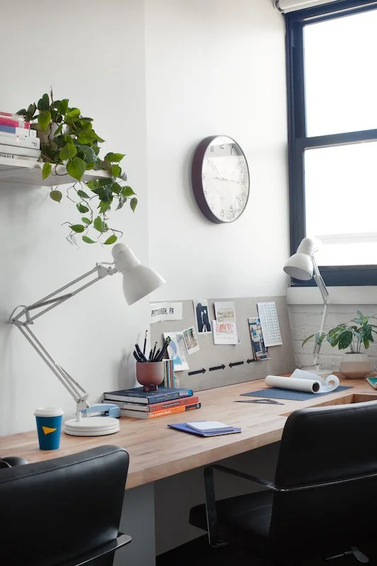 Office workspace with a white wall, two adjustable desk lamps, a shelf with a plant and books, a round wall clock, a large window, and a desk with notebooks, pens, a roll of paper, and potted plants.