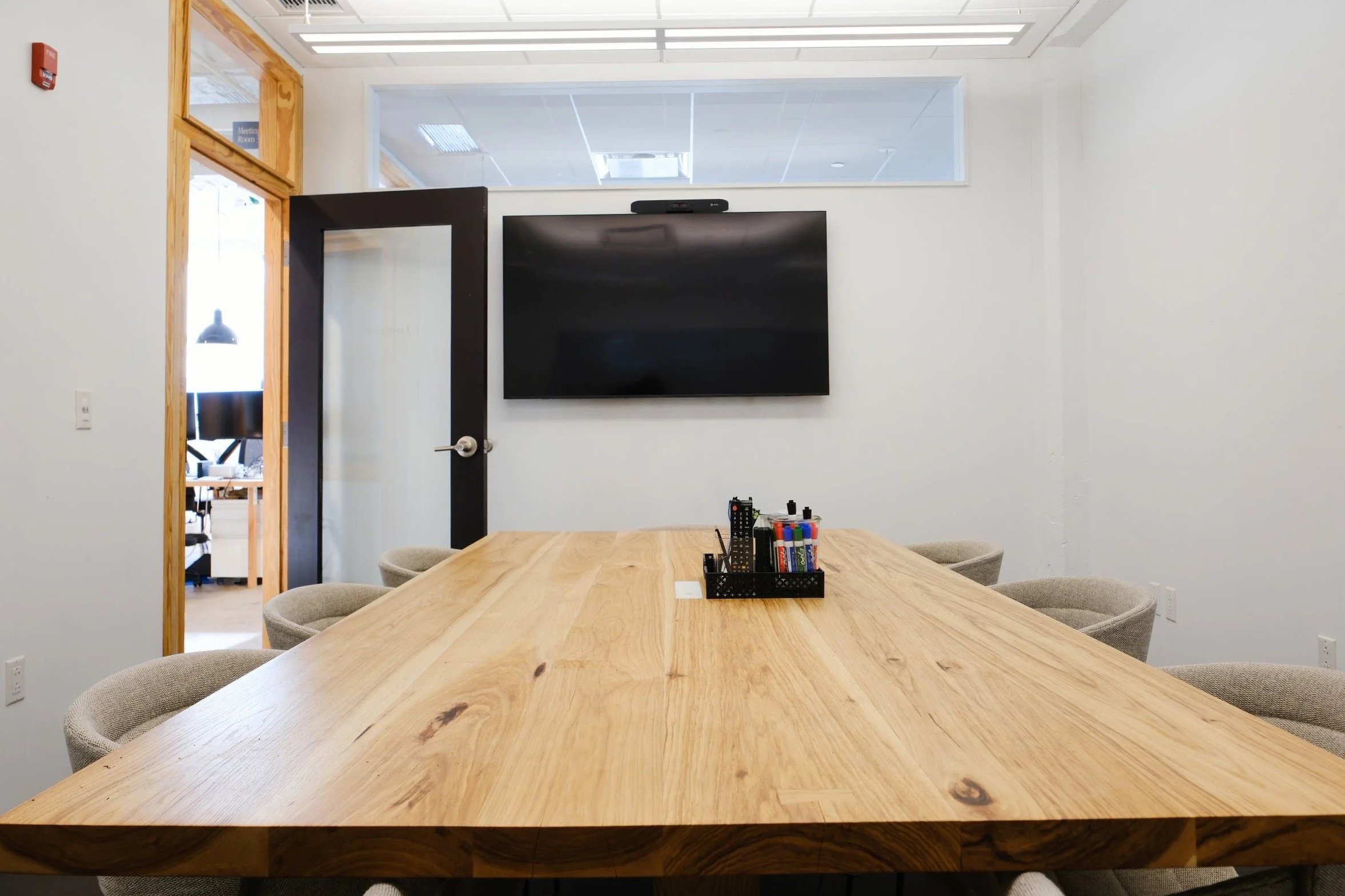 Spacious conference room with a large wooden table, six cushioned chairs, a wall-mounted TV, and a small container of markers in the center of the table.