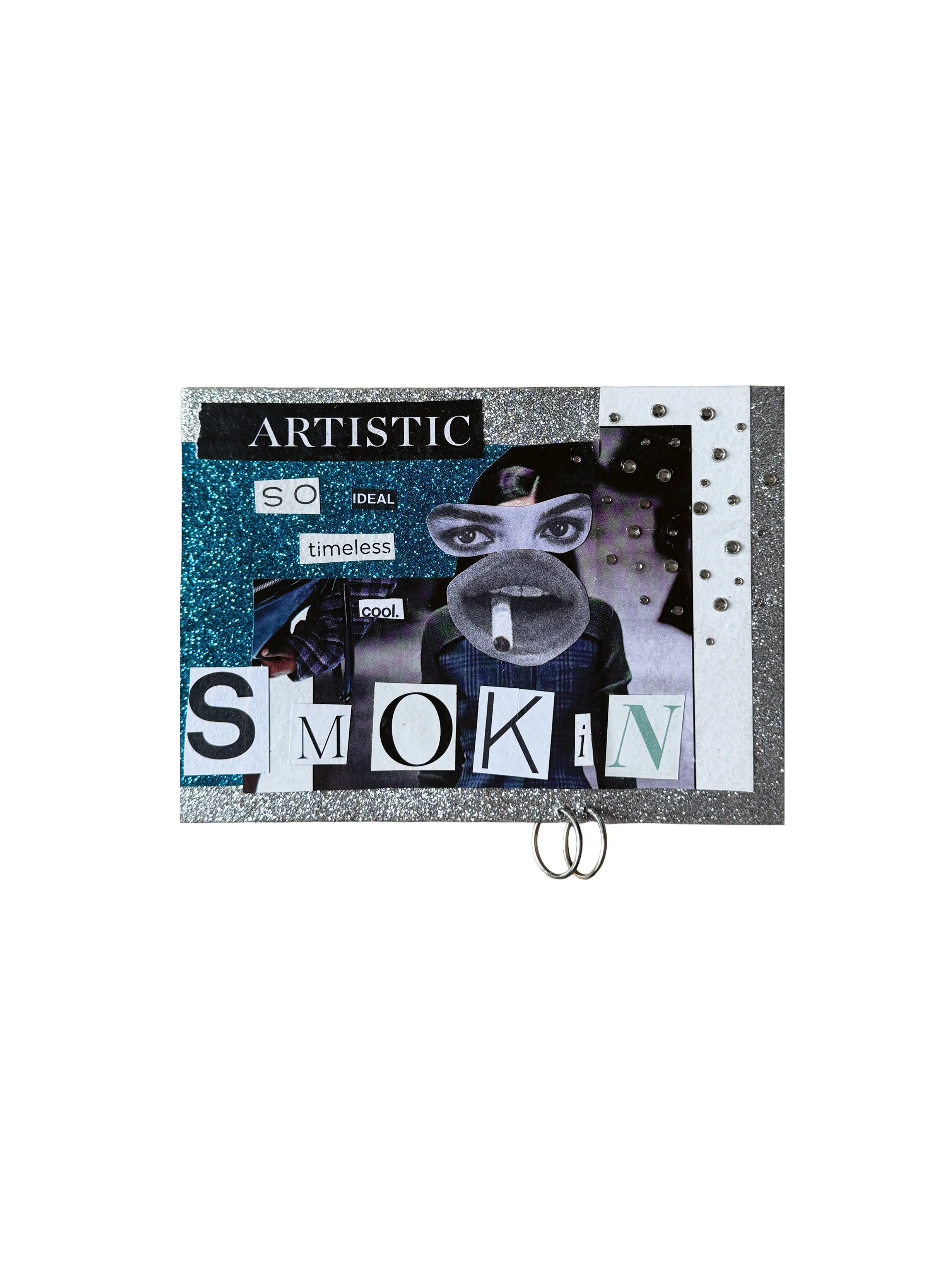 A collage-style art board with a glittery background, featuring a person's face wearing a mask, surrounded by words and cut-out letters including 'ARTISTIC', 'SO', 'IDEAL', 'timeless', 'cool', and 'SMOKIN'. The art board has metallic studs along one 