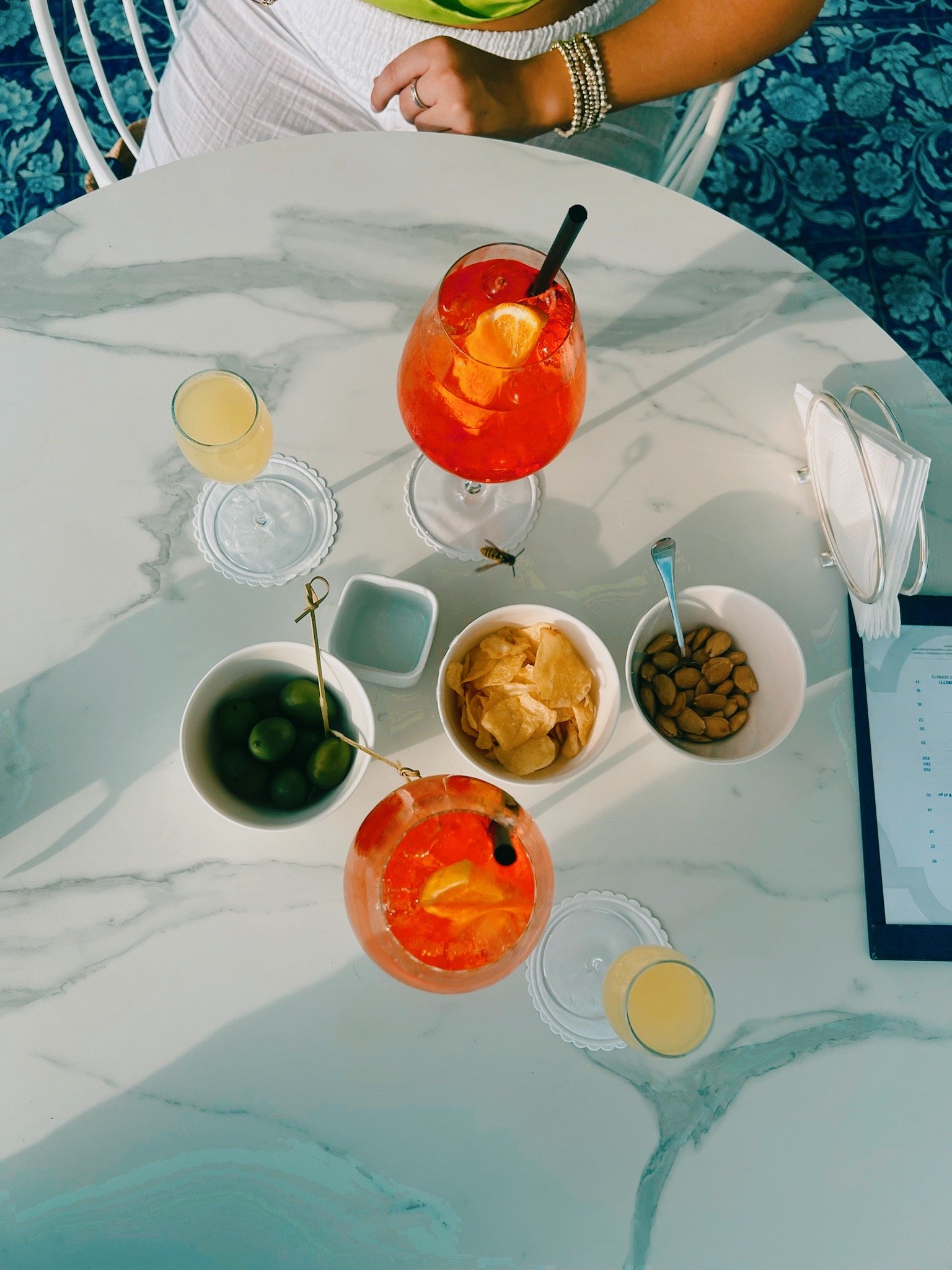 A white marble table with various drinks and snacks. Two glasses of orange-colored beverages, one garnished with an orange slice and a black straw, are on the table. There are bowls of green olives, potato chips, and mixed nuts. A person wearing bracelets and a ring is sitting at the table, with part of their arm and white skirt visible. The background includes a blue patterned wall and chairs.