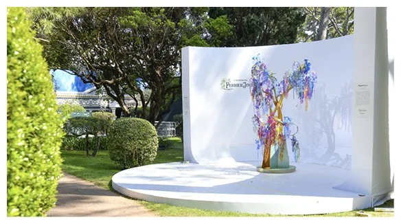 Une installation artistique composée d'un arbre en fleurs artificielles sur une scène arrondie en plein air, avec arbres et arbustes verts en arrière-plan.