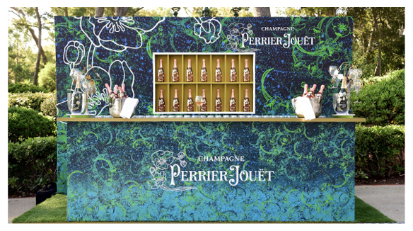 Un stand avec des bouteilles de champagne Perrier-Jouët, décoré avec un motif floral bleu et vert, situé en plein air dans un parc avec des arbres et des buissons.