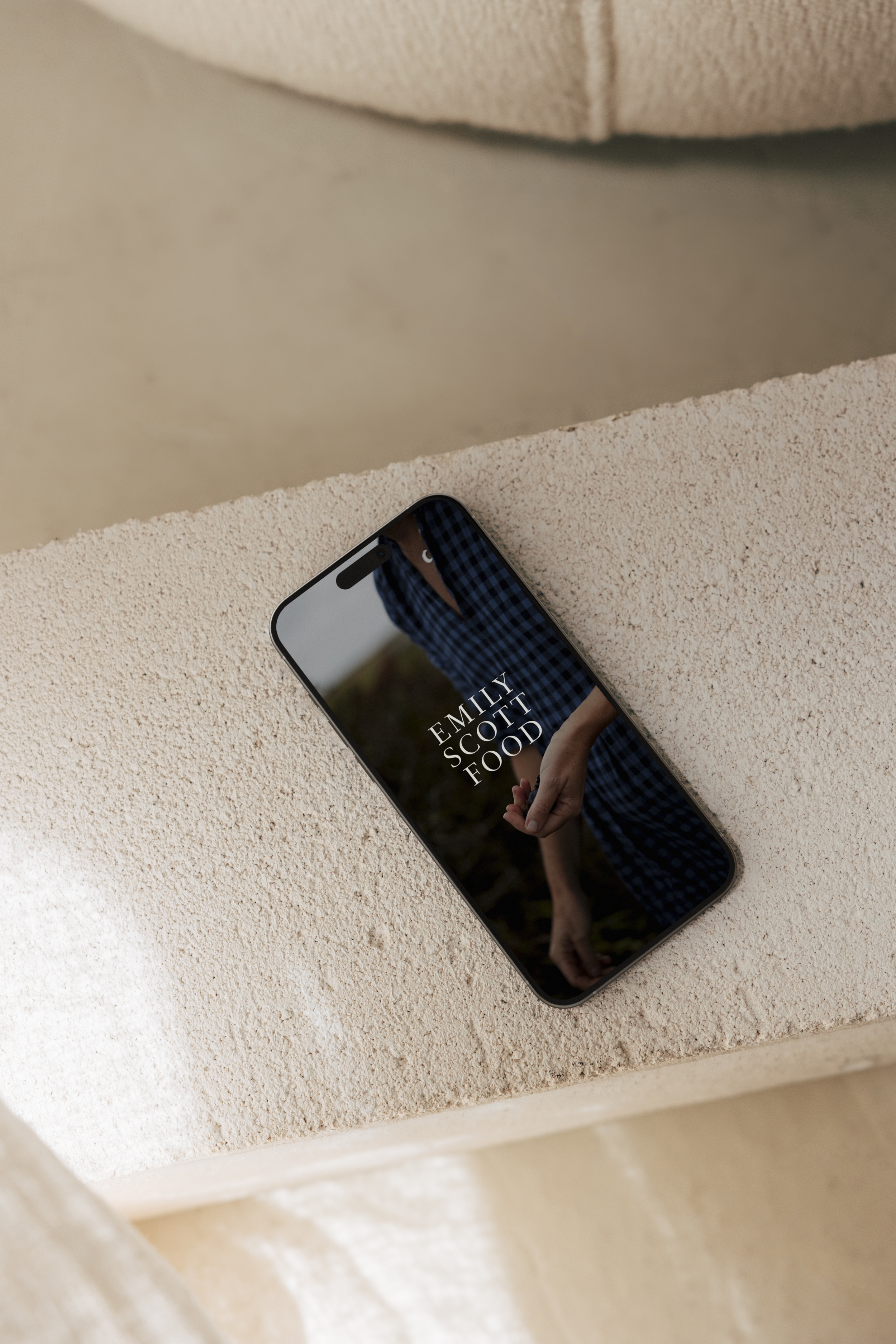 A smartphone with a black frame resting on a beige textured surface, displaying a book cover titled 'Emily Scott Food' with a person's arm and hand holding a small object.