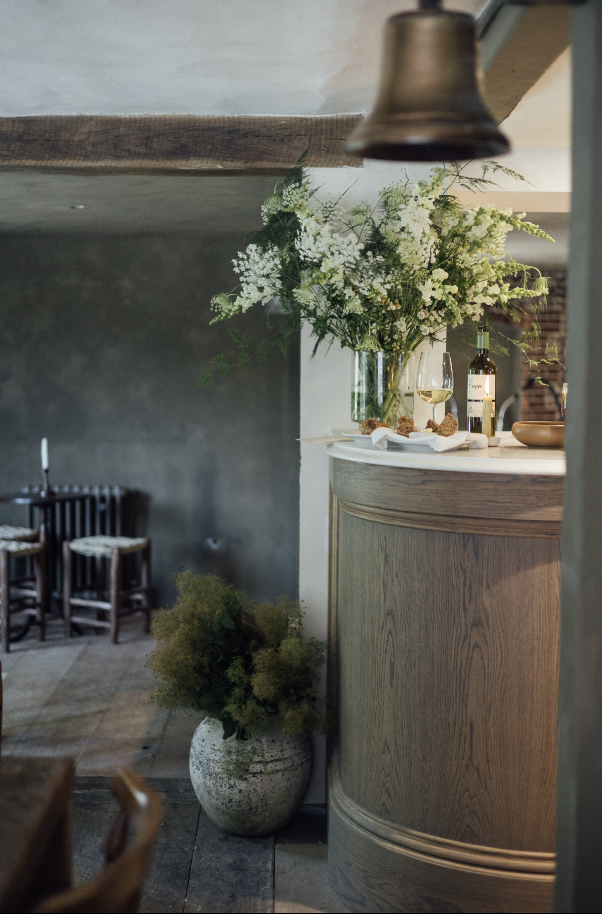 A floral arrangement with white flowers and greenery on a curved wooden counter, a glass of white wine and a bottle of wine beside it, with a small bowl and some bread on a white napkin. In the background, there are chairs along a wall and a dark textured wall.