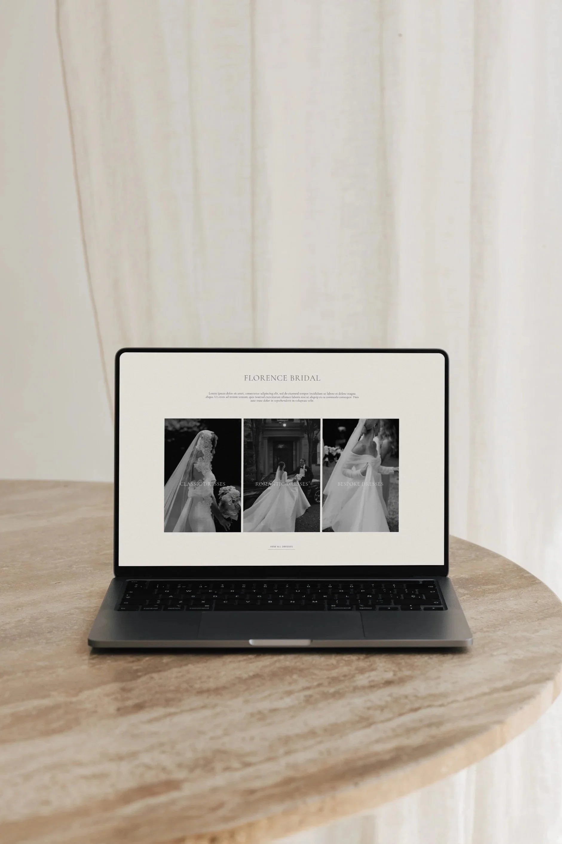 Laptop on a beige marble table displaying a wedding dress website with black and white photos of brides.
