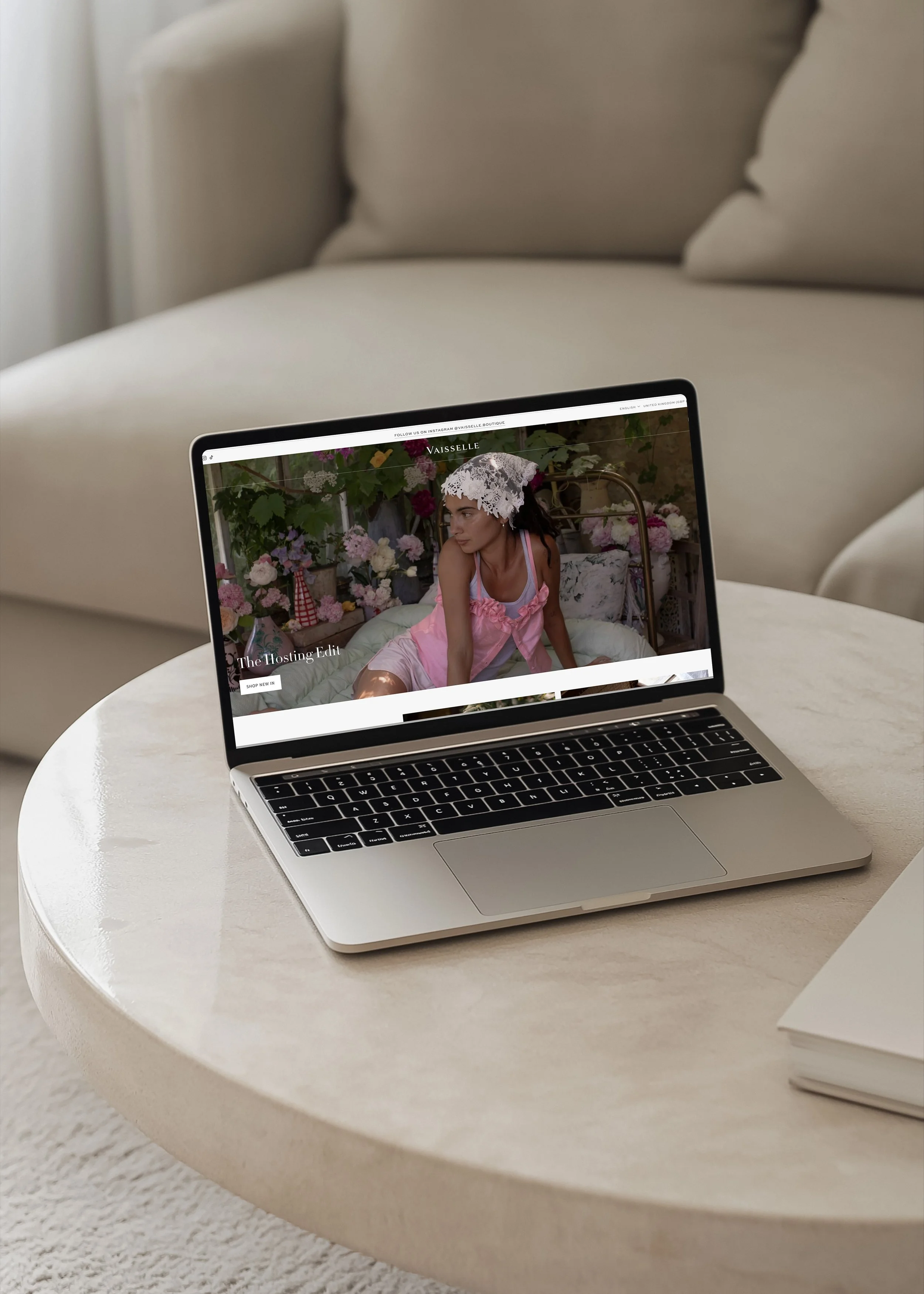 A laptop on a white round table showing a fashion website with a woman in pink lingerie and lace headscarf surrounded by flowers.