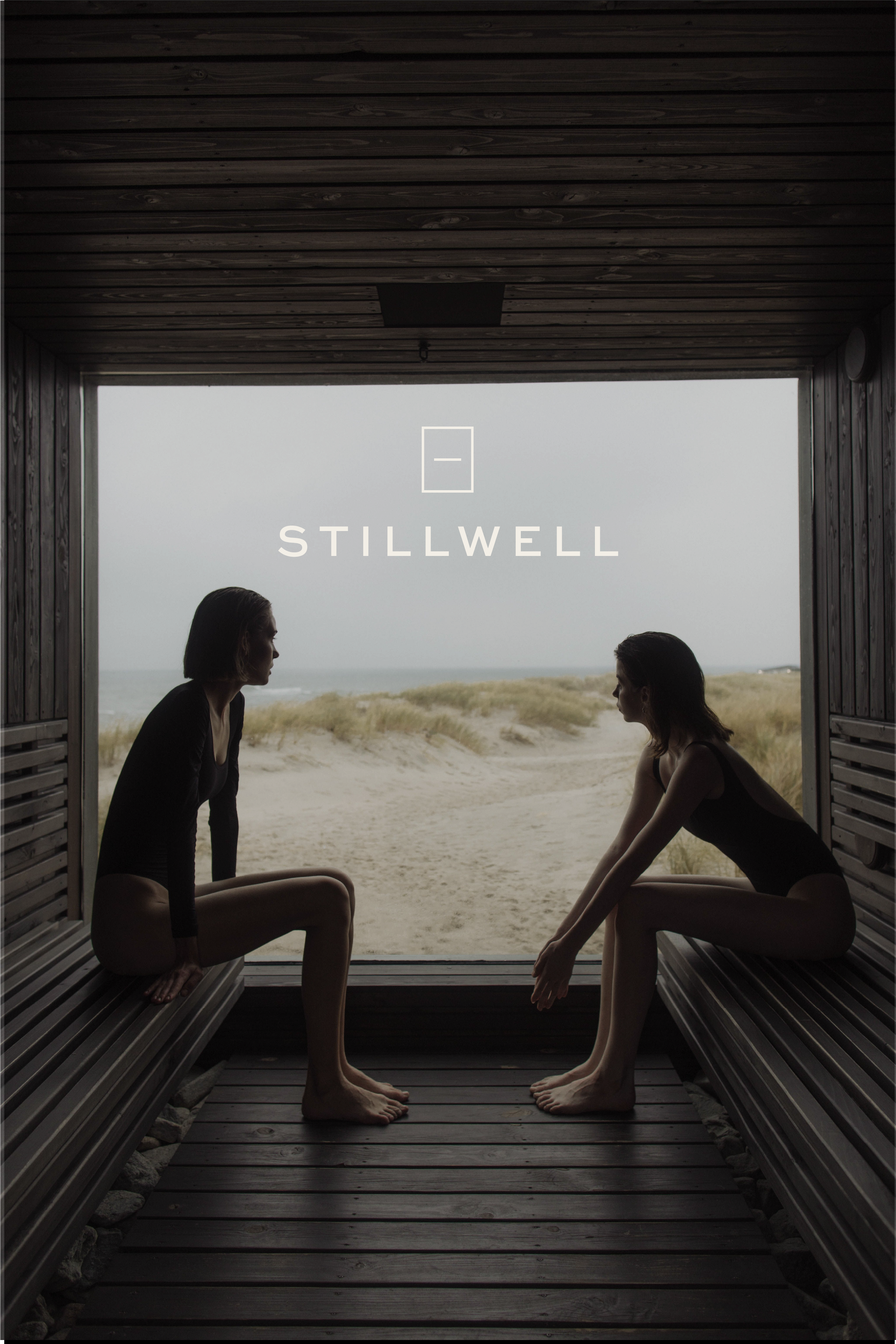 Two women practicing yoga in a wooden beach cabana facing outside towards the sea, sitting in a mirror image pose with knees bent, one hand on the ground, and the other hand on their knee, with large windows showing sandy dunes and cloudy sky.