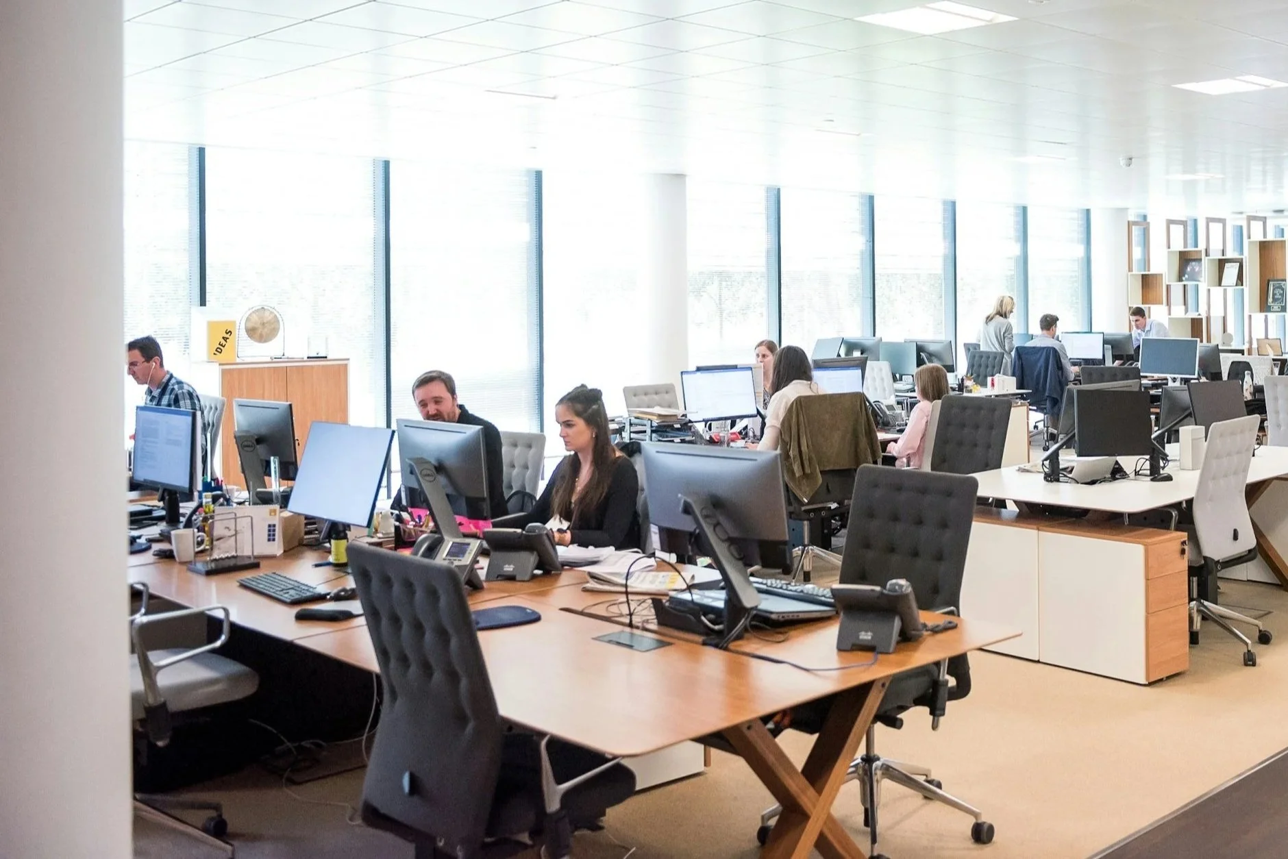 A modern open-plan office with multiple employees working at their desks, multiple computer monitors, and large windows providing natural light.