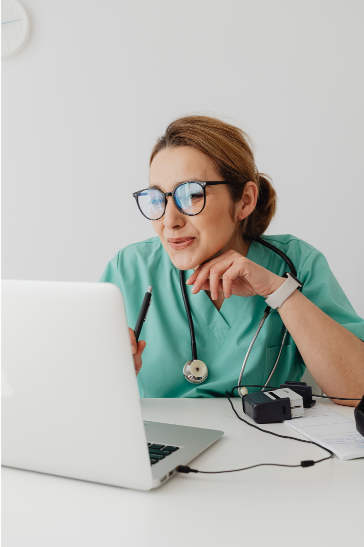 A female healthcare professional in scrubs with glasses and a stethoscope around her neck, looking confused and holding a stylus while looking at a laptop.