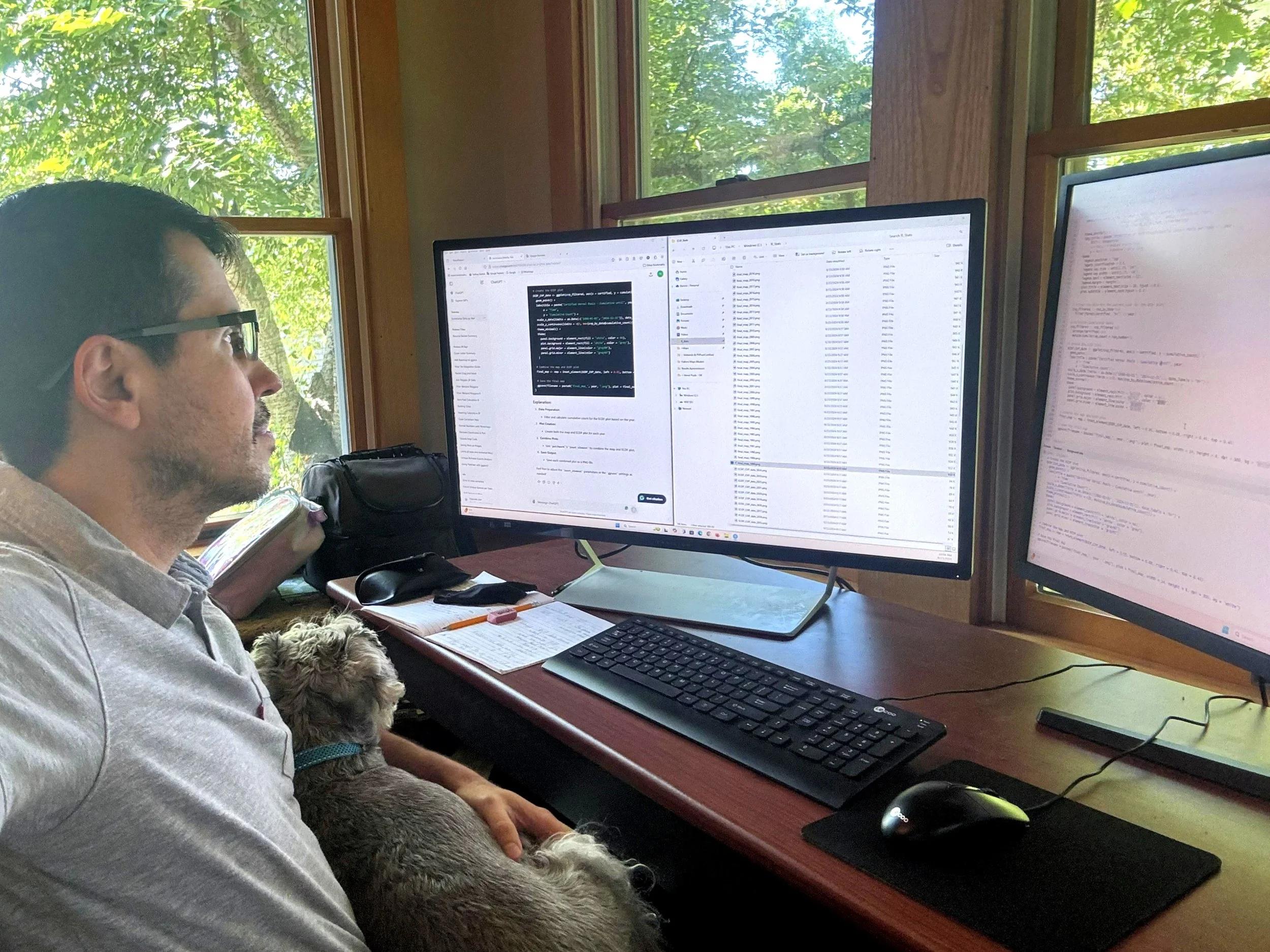 A man working at a desk with two monitors, one displaying coding and data tables, the other displaying maps, with a dog sitting on his lap near a window showing trees outside.