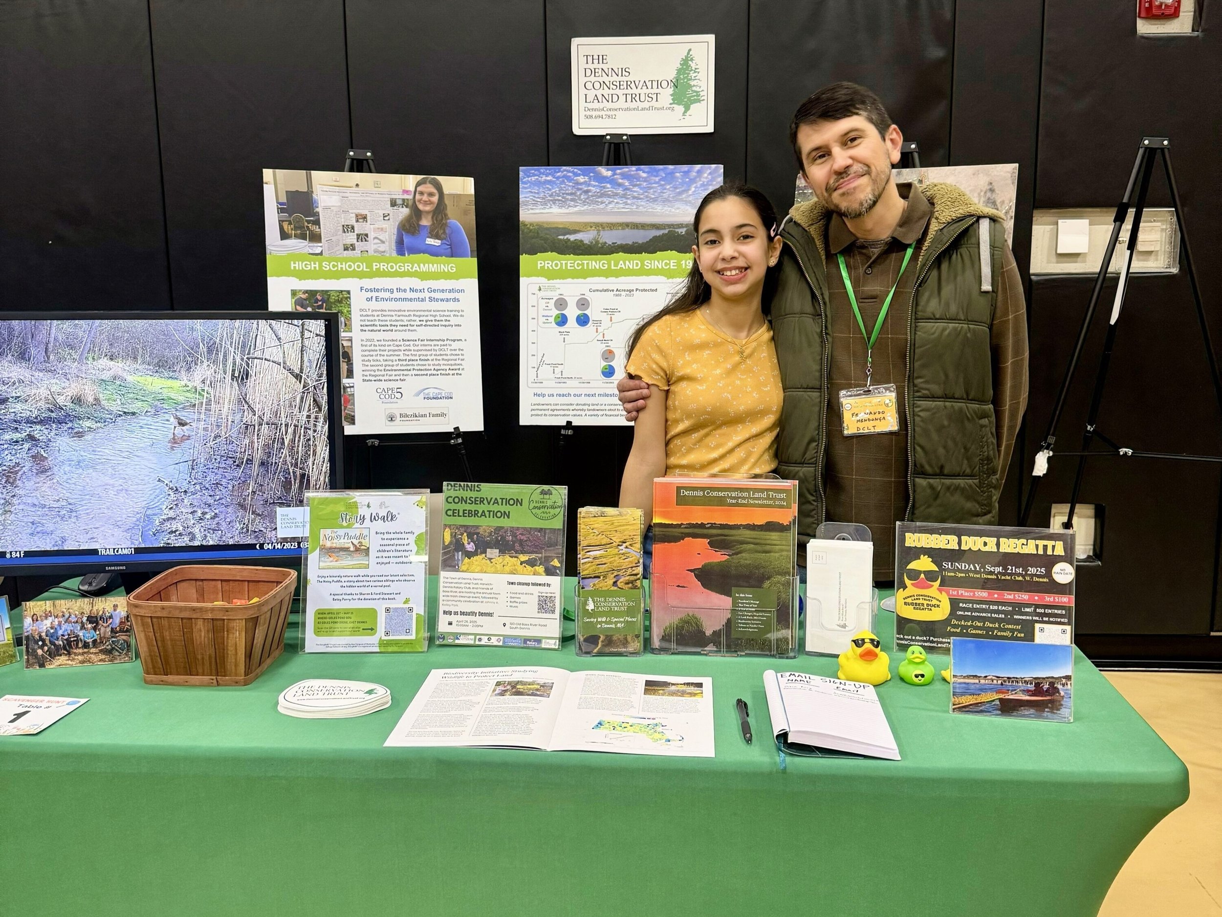 Two people, a young girl and a man, standing behind a green table at an environmental conservation event. The table displays brochures, flyers, a small photo, and rubber duck toys. The background features informational posters about land conservation, a monitor showing a nature scene, and a sign for the Dennis Conservation Land Trust.