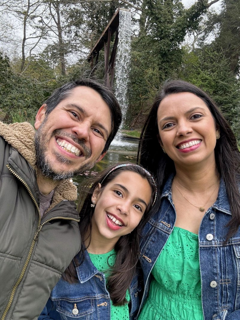 A family of three smiling and taking a selfie outdoors near a waterfall with trees in the background.