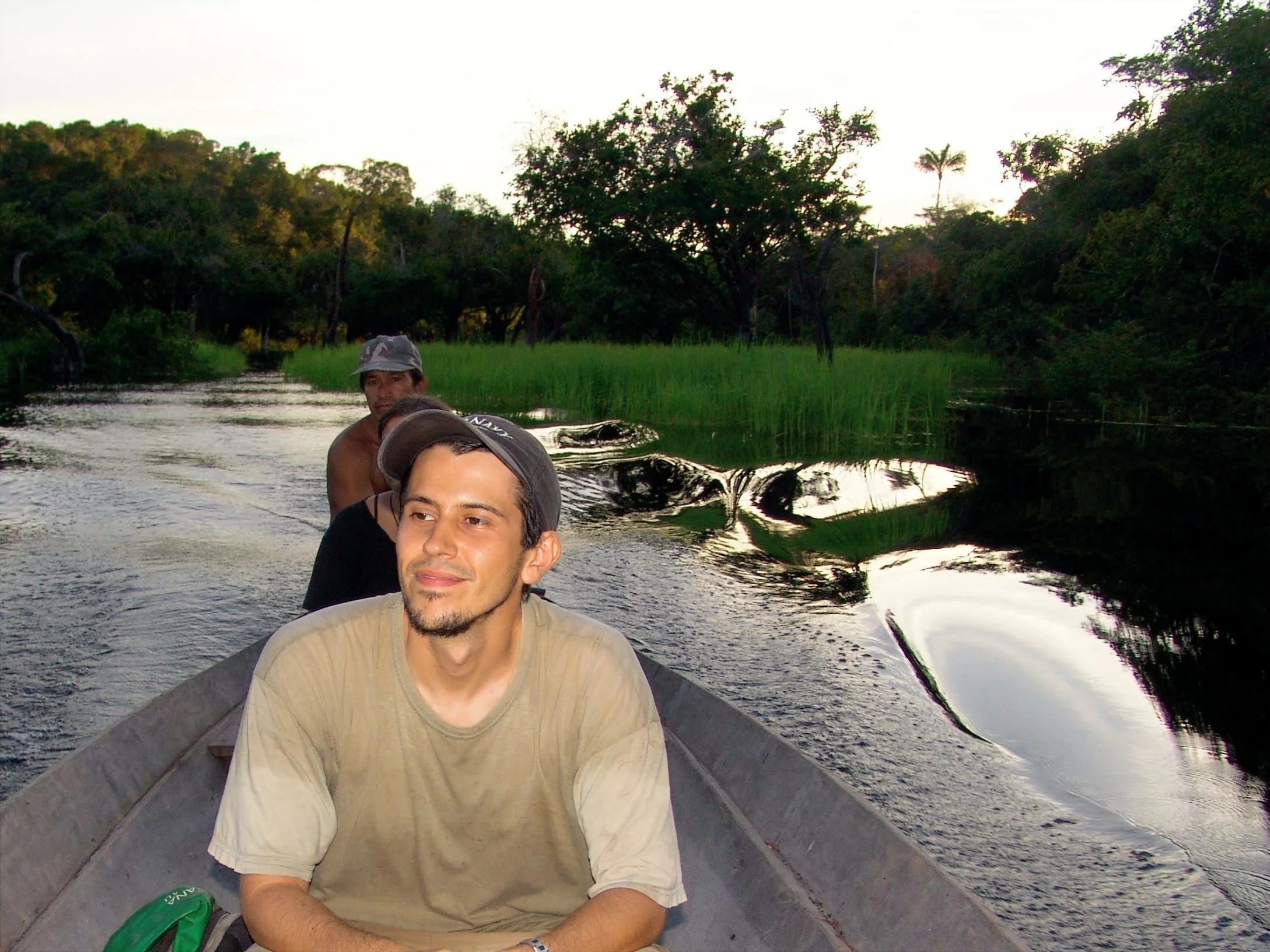 Three men are on a boat traveling through a river surrounded by lush green trees and tall grass, with a calm water surface reflecting the scene.