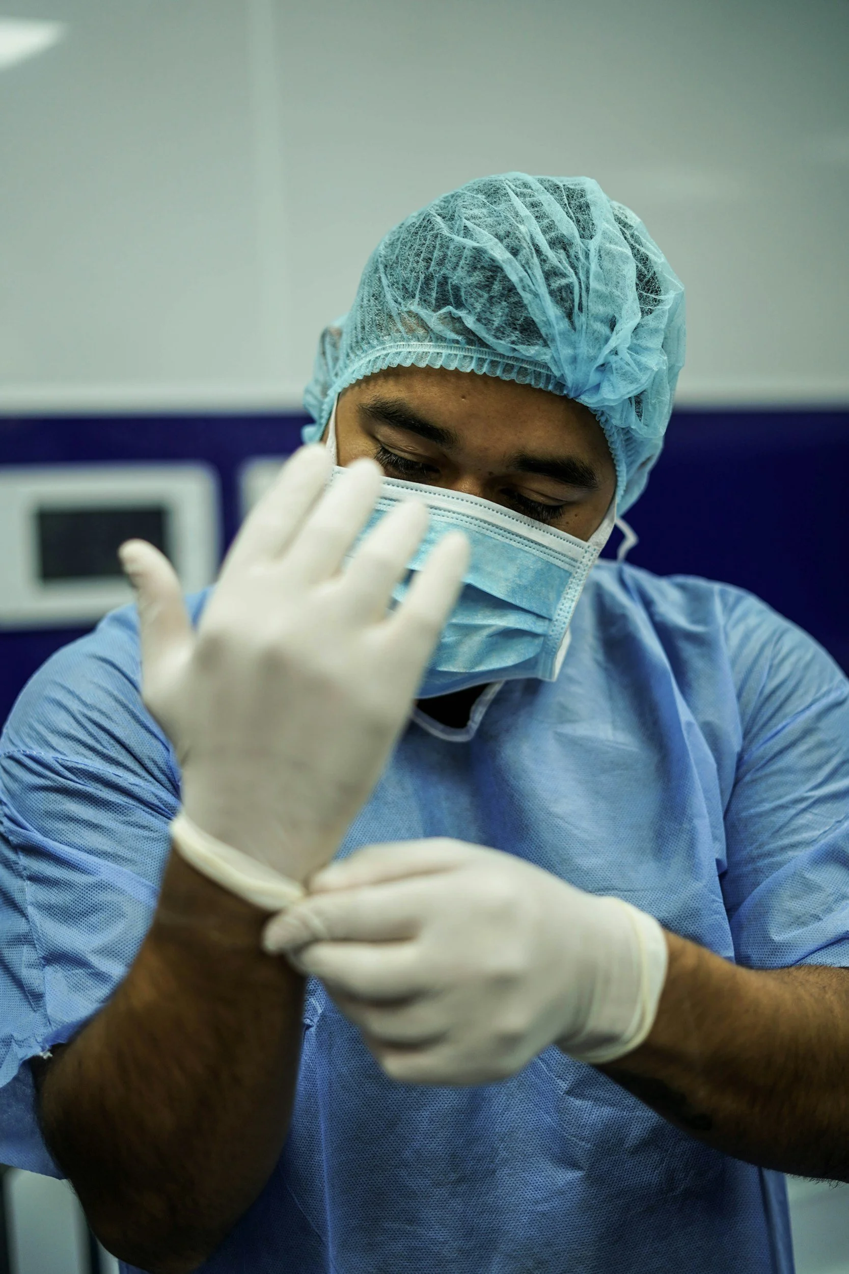 Medical professional in protective gown, hairnet, face mask, and gloves adjusting their glove in a clinical setting.