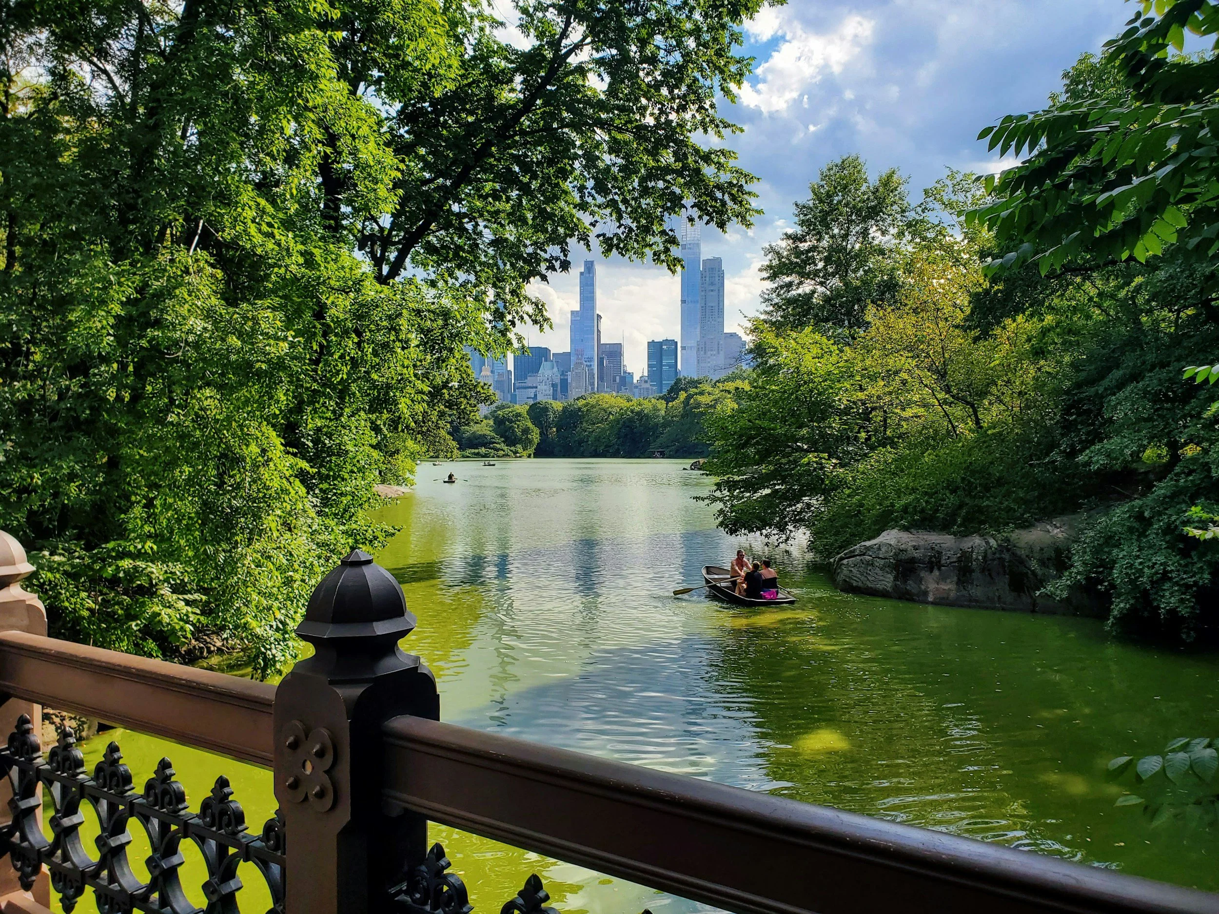 A view of a city skyline with tall buildings seen across a green lake, with people in small boats and trees on both sides, taken from a park with a decorative fence.