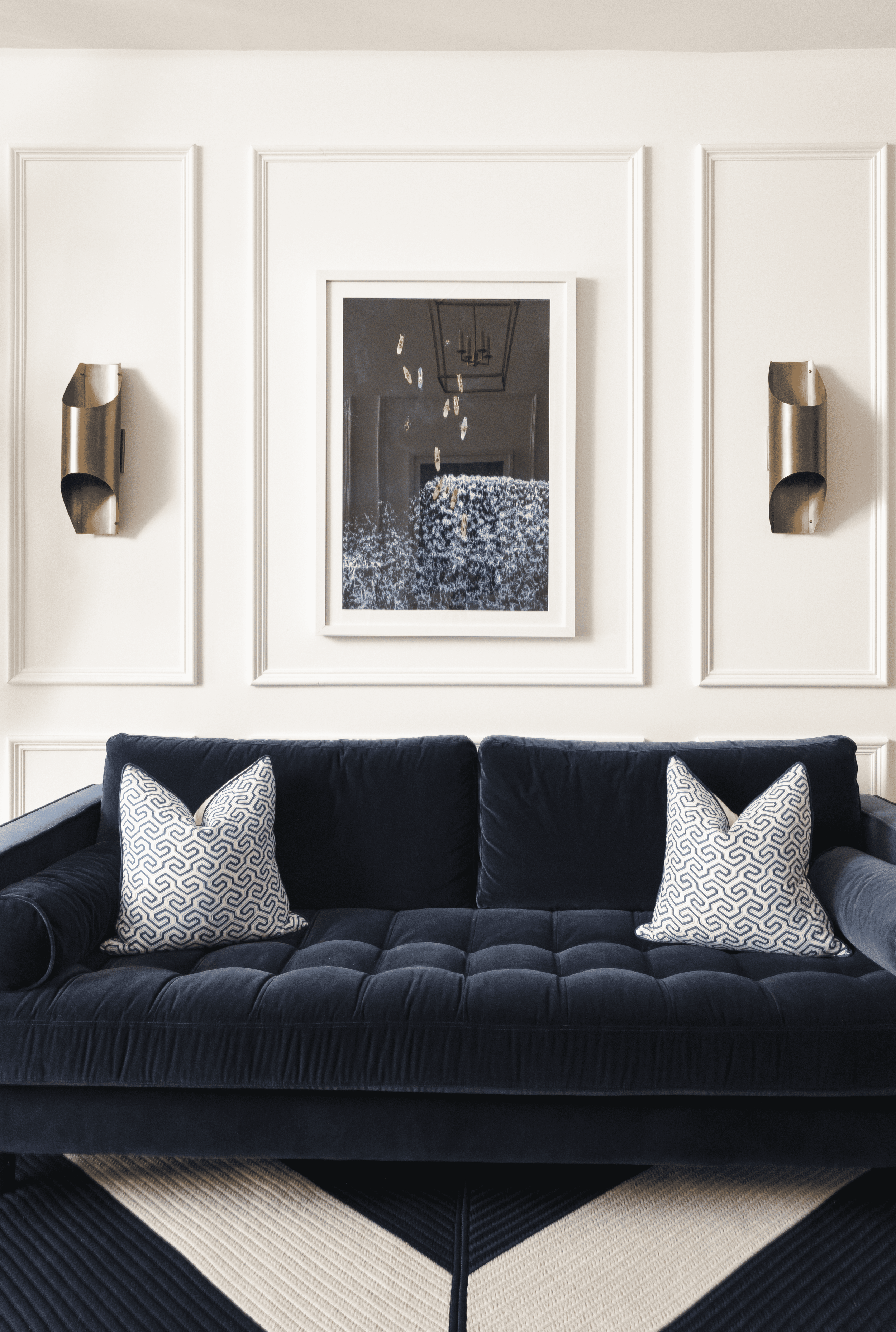 A navy blue velvet sofa with three decorative throw pillows containing a geometric pattern, situated on a black and white striped area rug in front of a white wall with decorative molding. A framed mirror and two metallic wall sconces are mounted on the wall.