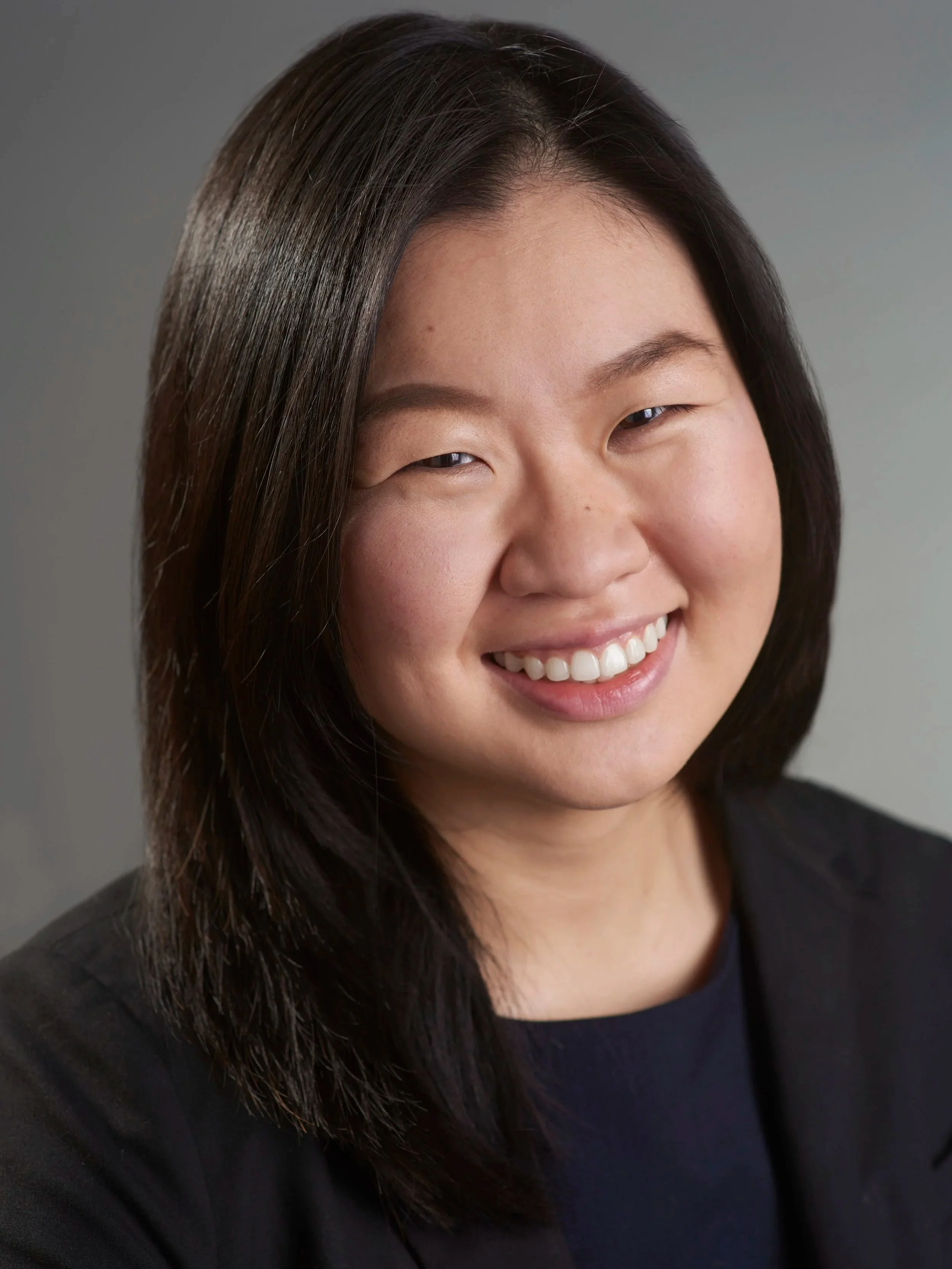 Close-up portrait of an Asian woman with shoulder-length dark hair smiling against a light gray background.