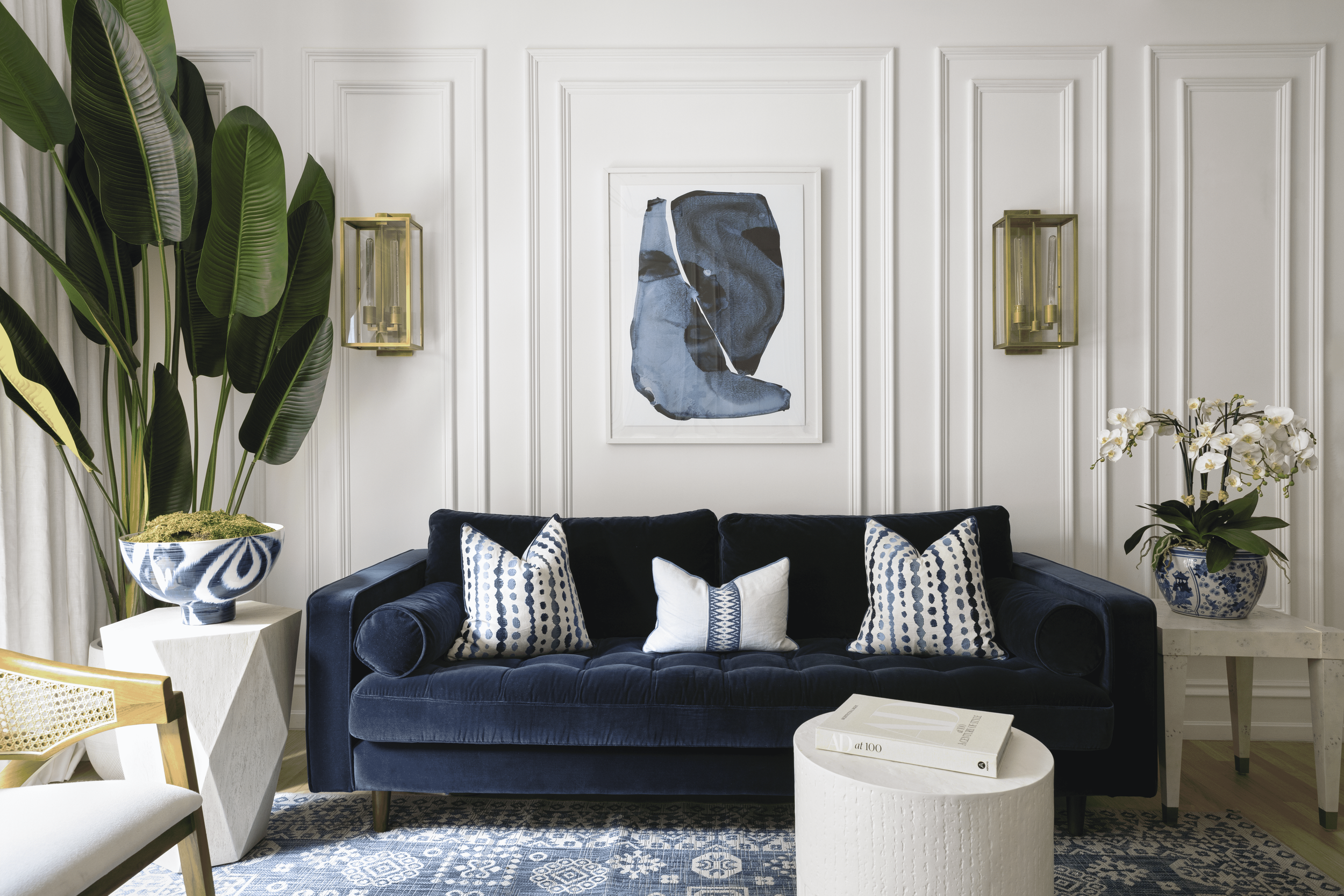 Living room with navy velvet sofa, decorative pillows, and artwork on white paneled walls. Large potted plants on white side tables and orchid flowers. Photo taken by Trevor Parker