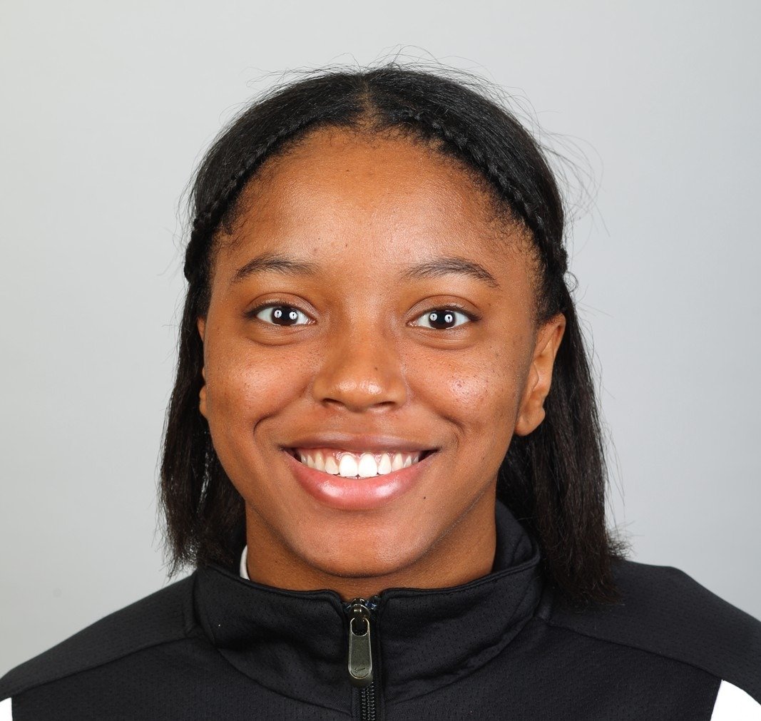 Headshot of a young woman with a bright smile, wearing a black zip-up jacket, against a plain light gray background.