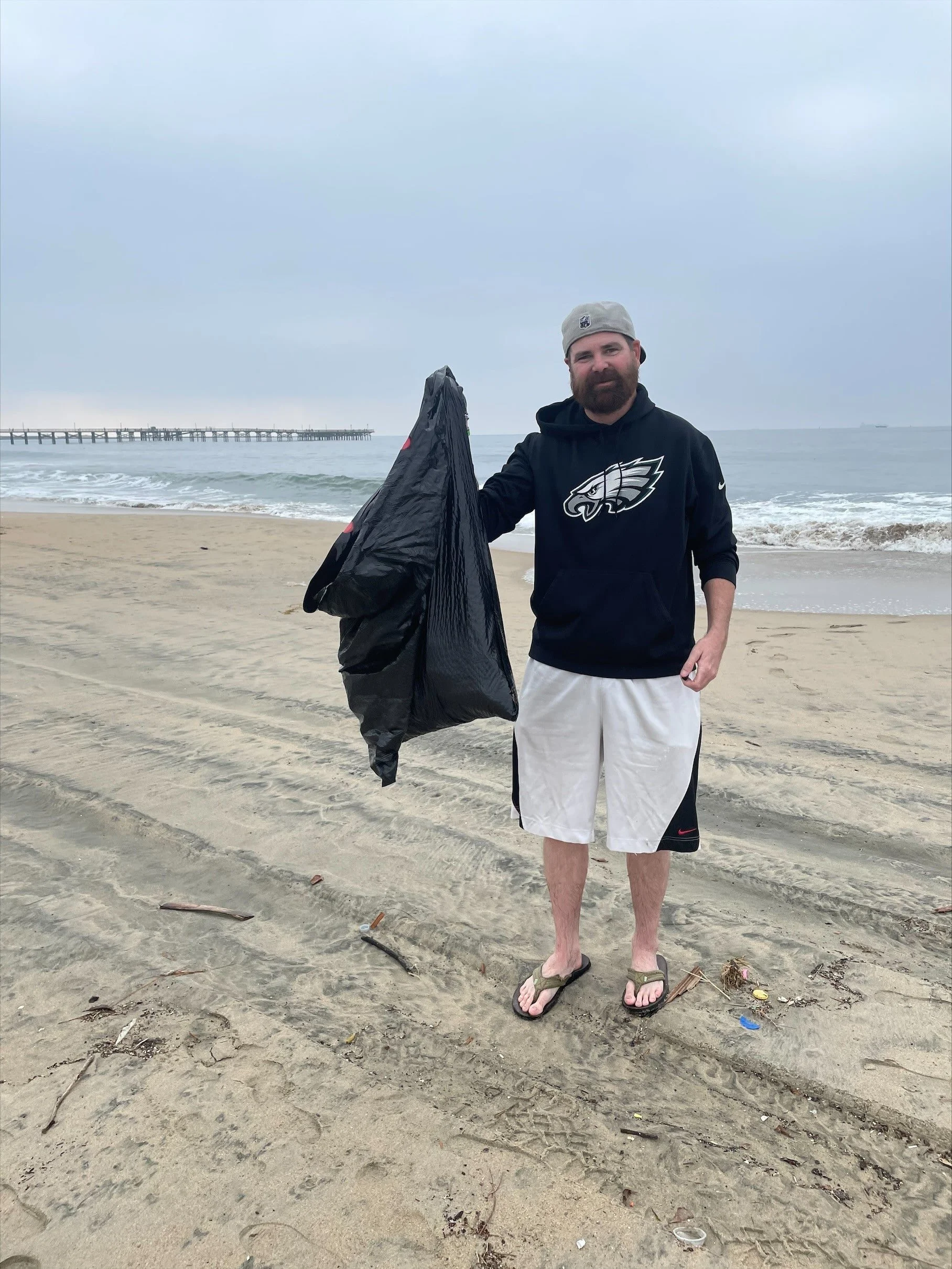 A man with a beard wearing a gray baseball cap, black hoodie with a sports team logo, white shorts, and sandals, standing on a sandy beach holding a large black trash bag. In the background, the ocean, a pier, and an overcast sky are visible.