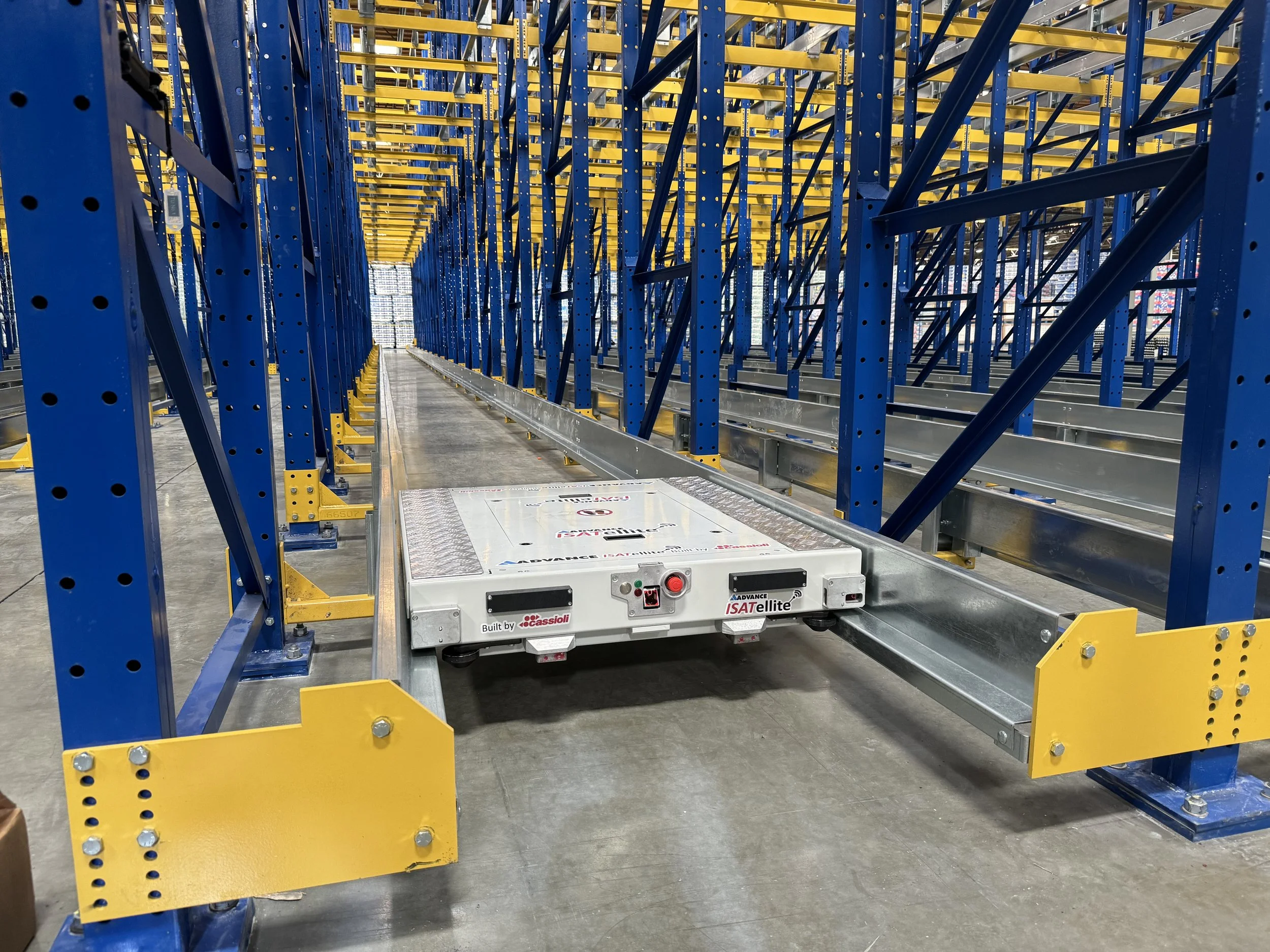 A warehouse storage area with blue and yellow metal shelving racks and an autonomous robot on the floor labeled 'Advance ISATEllite' used for moving items among the shelves.