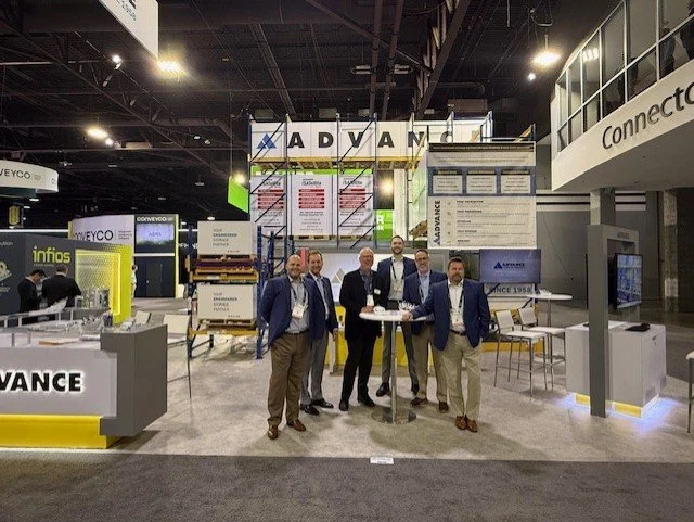 Five men in business attire standing and posing for a photo at a trade show booth with giant signs displaying the word "ADVANCE" in the background.
