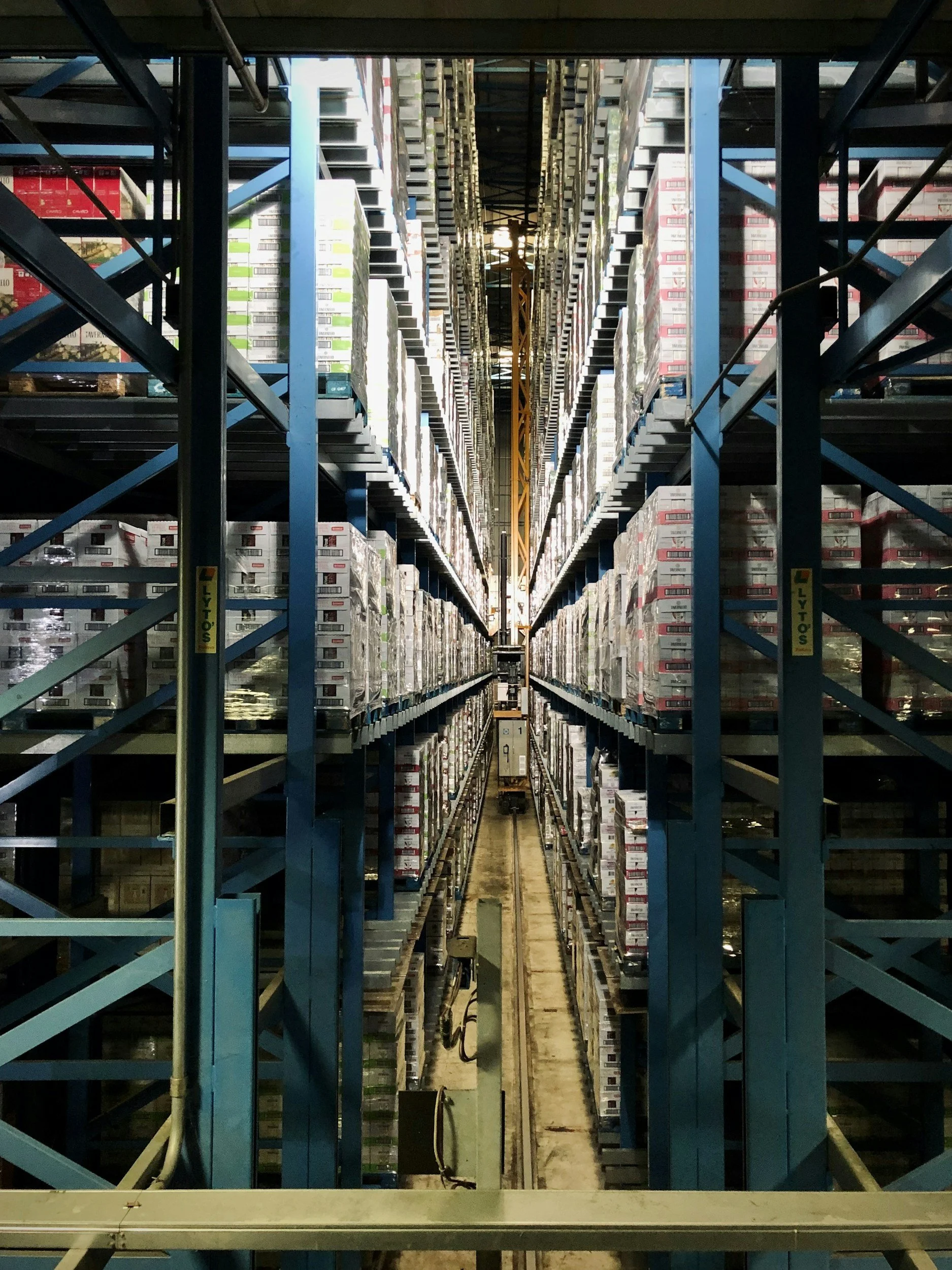 Multiple levels of warehouse shelving filled with boxes and a crane in the background.