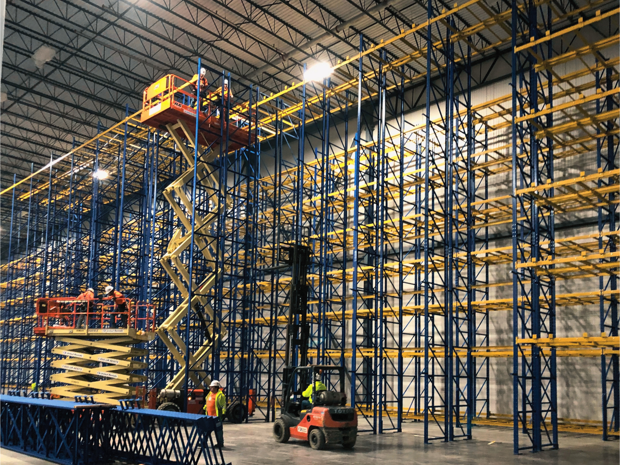 Workers in bright vests and scissor-lifts install pallet racking in a warehouse.