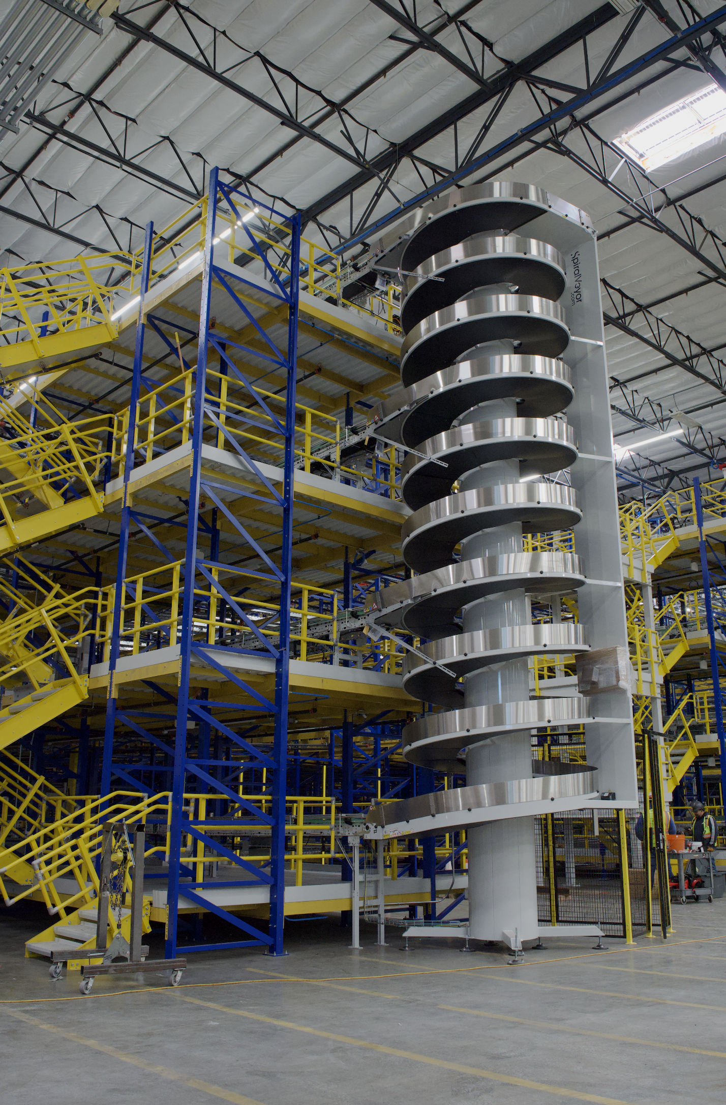 Inside a large industrial warehouse with a metal spiral staircase painted yellow, steel framework, and some machinery including a large metallic spiral structure.