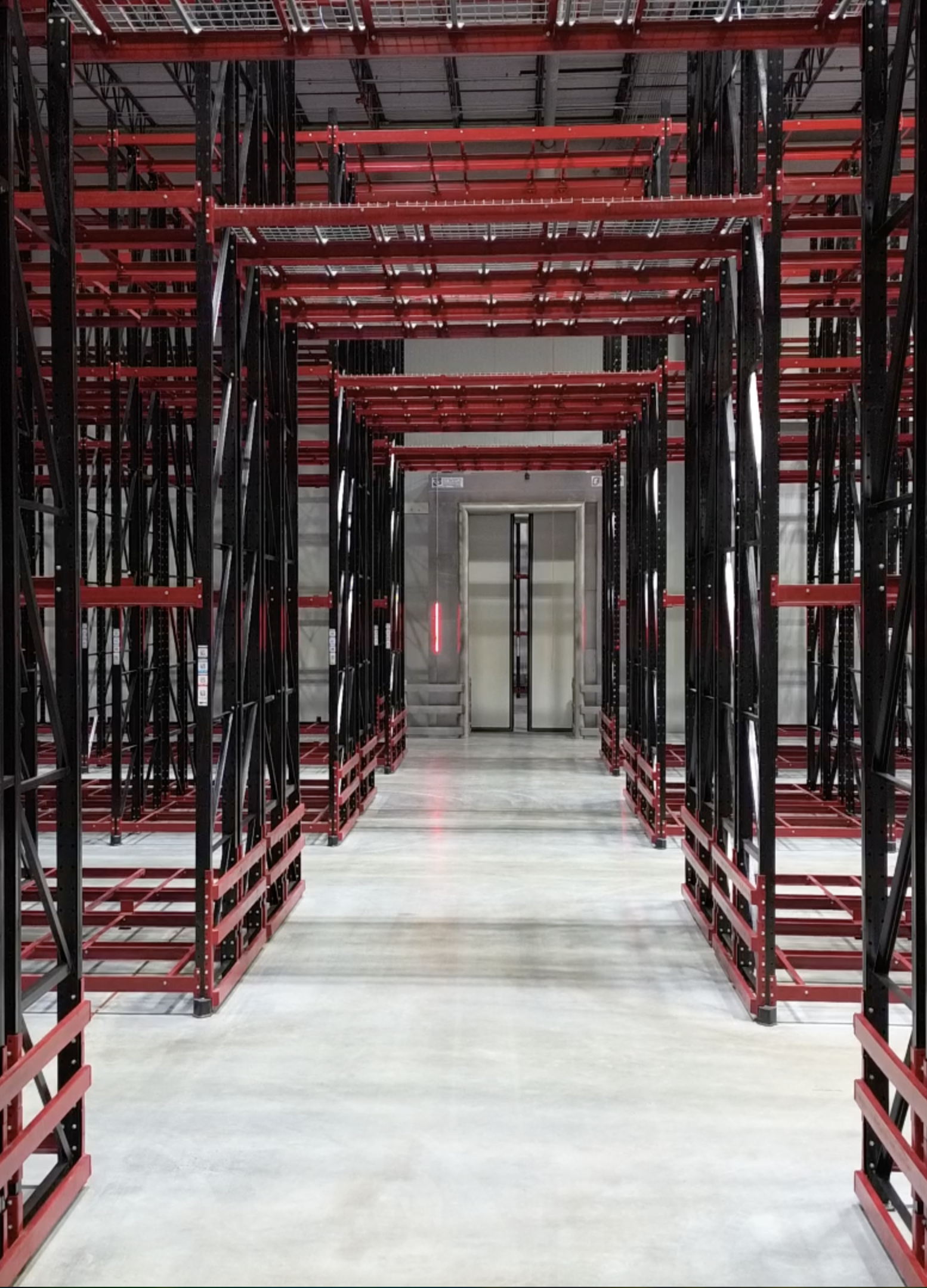 Empty industrial storage racks with red beams and black supports in a warehouse, with an elevator in the background.