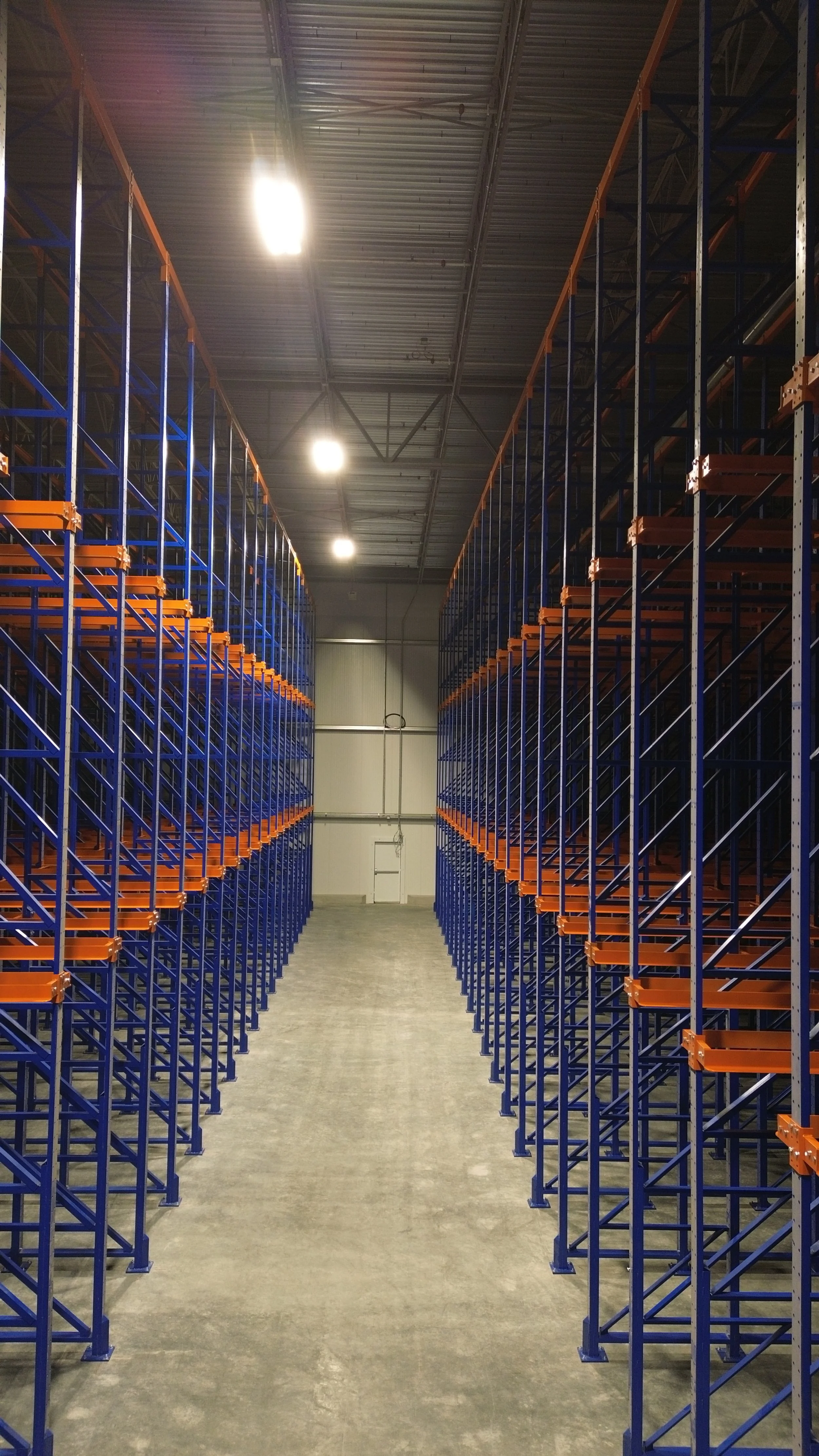Empty warehouse aisle with tall blue and orange metal shelving racks on both sides, brightly lit ceiling, and gray concrete floor.