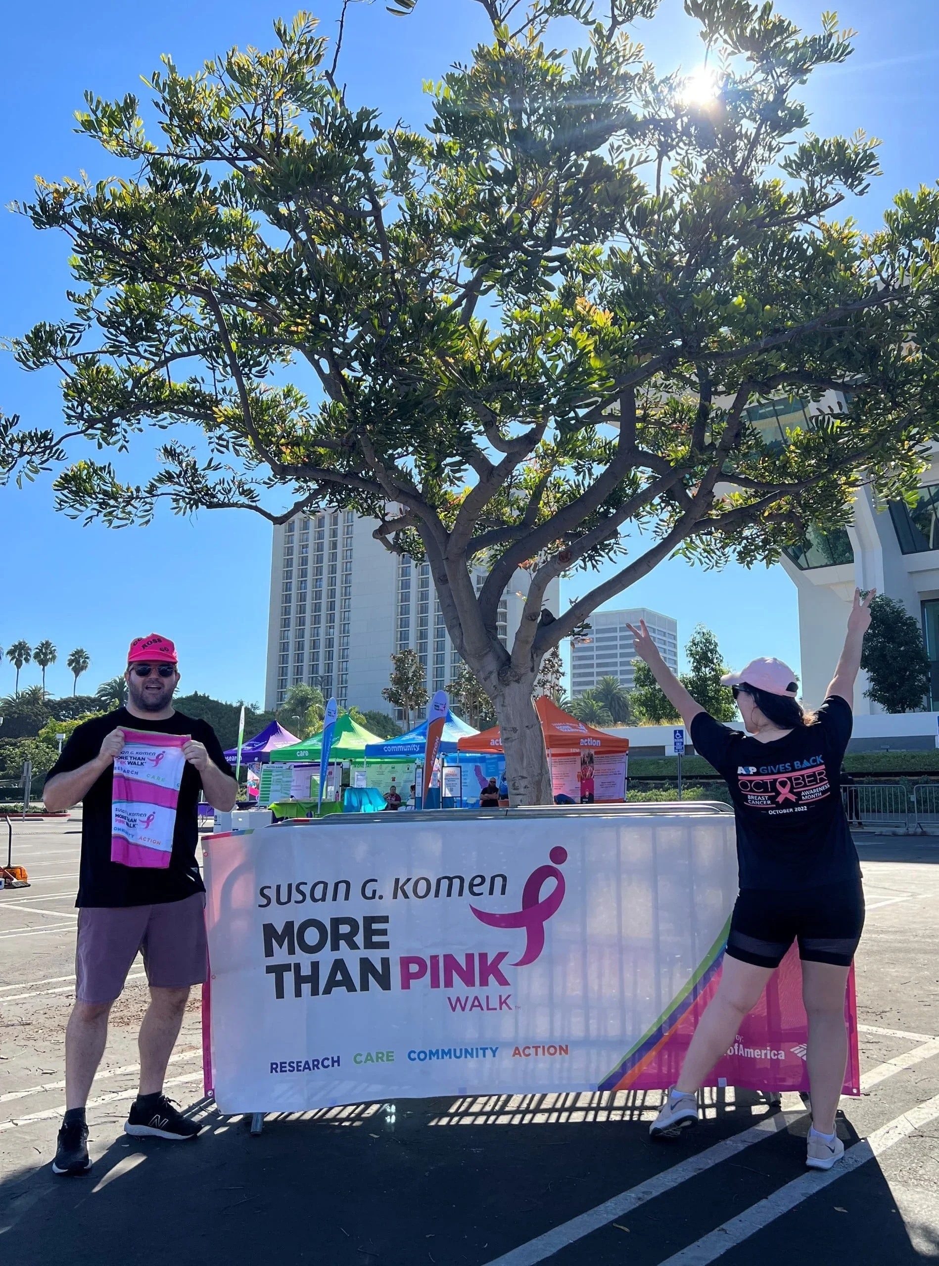 Two people standing outdoors behind a pink and white banner for the Susan G. Komen More Than Pink Walk, with a large tree, buildings, and blue sky in the background. One person is holding a race bib, and the other has their arms raised.