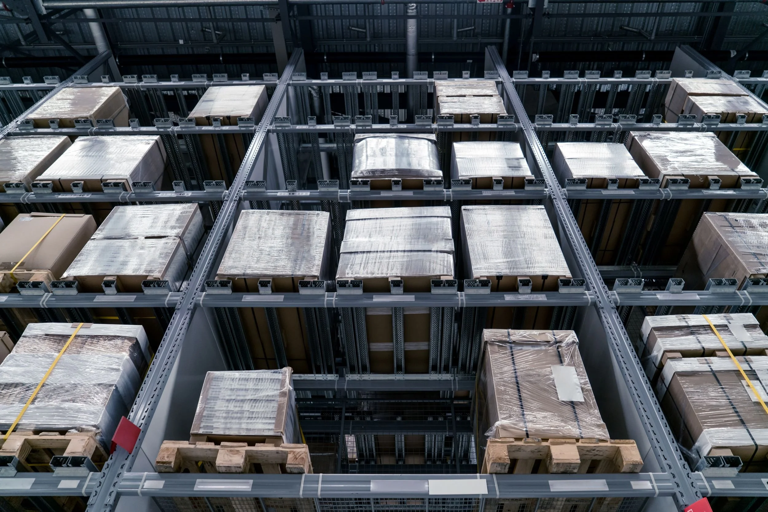 Warehouse storage racks filled with cardboard boxes wrapped in plastic, stacked neatly on wooden pallets.