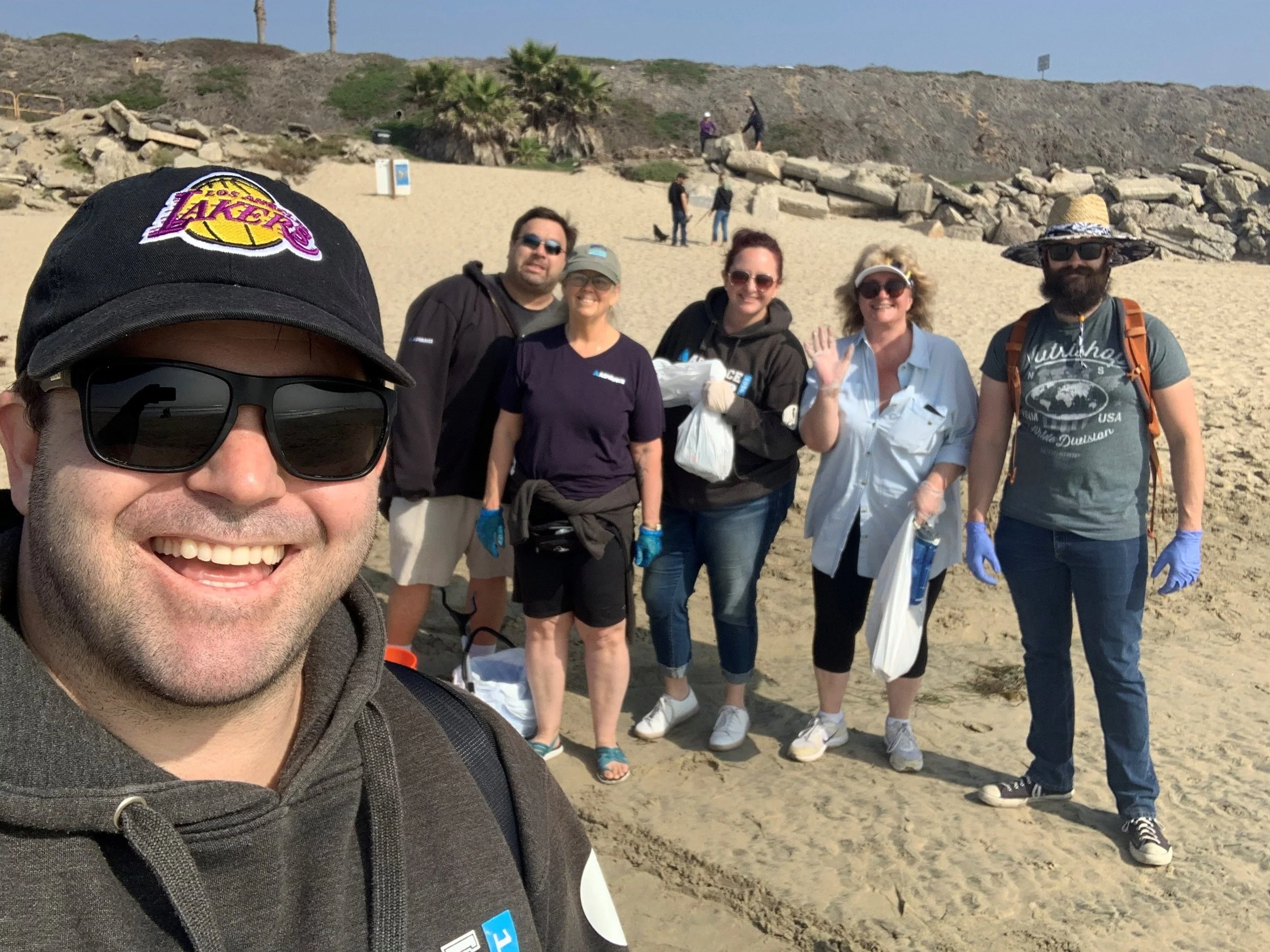A group of people standing together on a sandy beach, smiling and waving at the camera, with a rocky hillside and palm trees in the background.