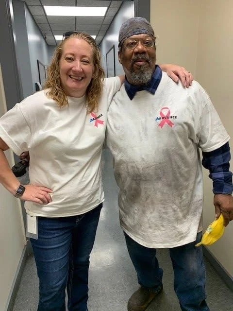 Two people, a woman and a man, standing in a hallway with their arms around each other. Both are wearing white T-shirts with pink ribbons and the word 'ADVANCE' printed on them, indicating support for breast cancer awareness. The woman has curly blonde hair, and the man is wearing a beanie and has a beard.