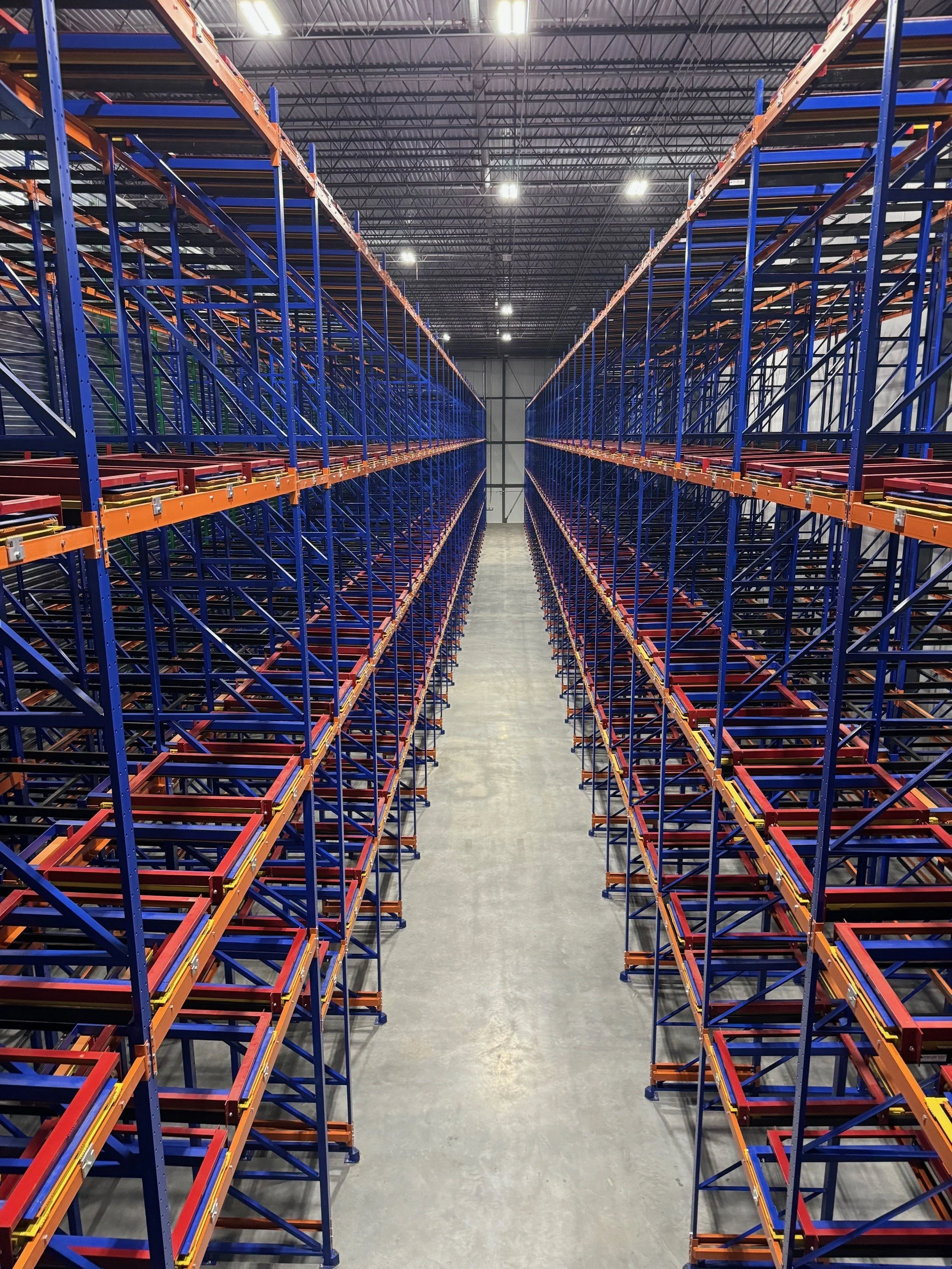 Empty warehouse with tall blue and orange shelves extending into the distance, no products visible, concrete floor, illuminated ceiling