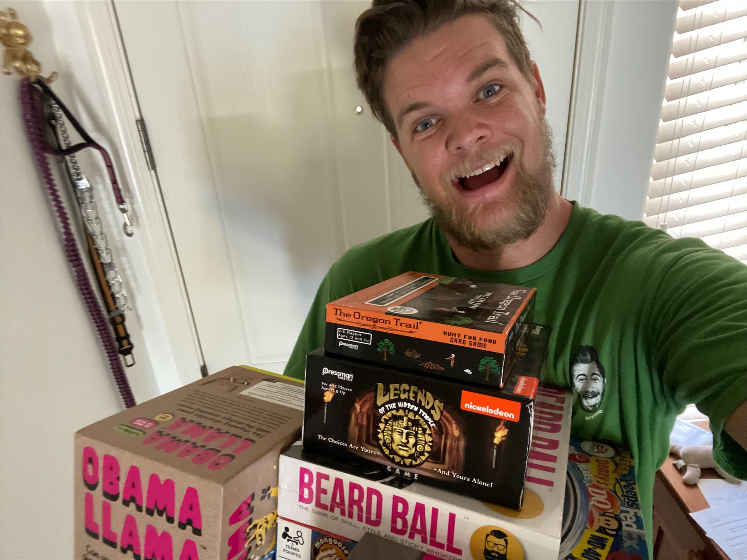 A man with a beard and short hair wearing a green shirt smiling happily and holding a stack of board games, including 'Beard Ball', 'Legends of the Hidden Temple', and 'The Oregon Trail', in a room with white walls and window blinds.